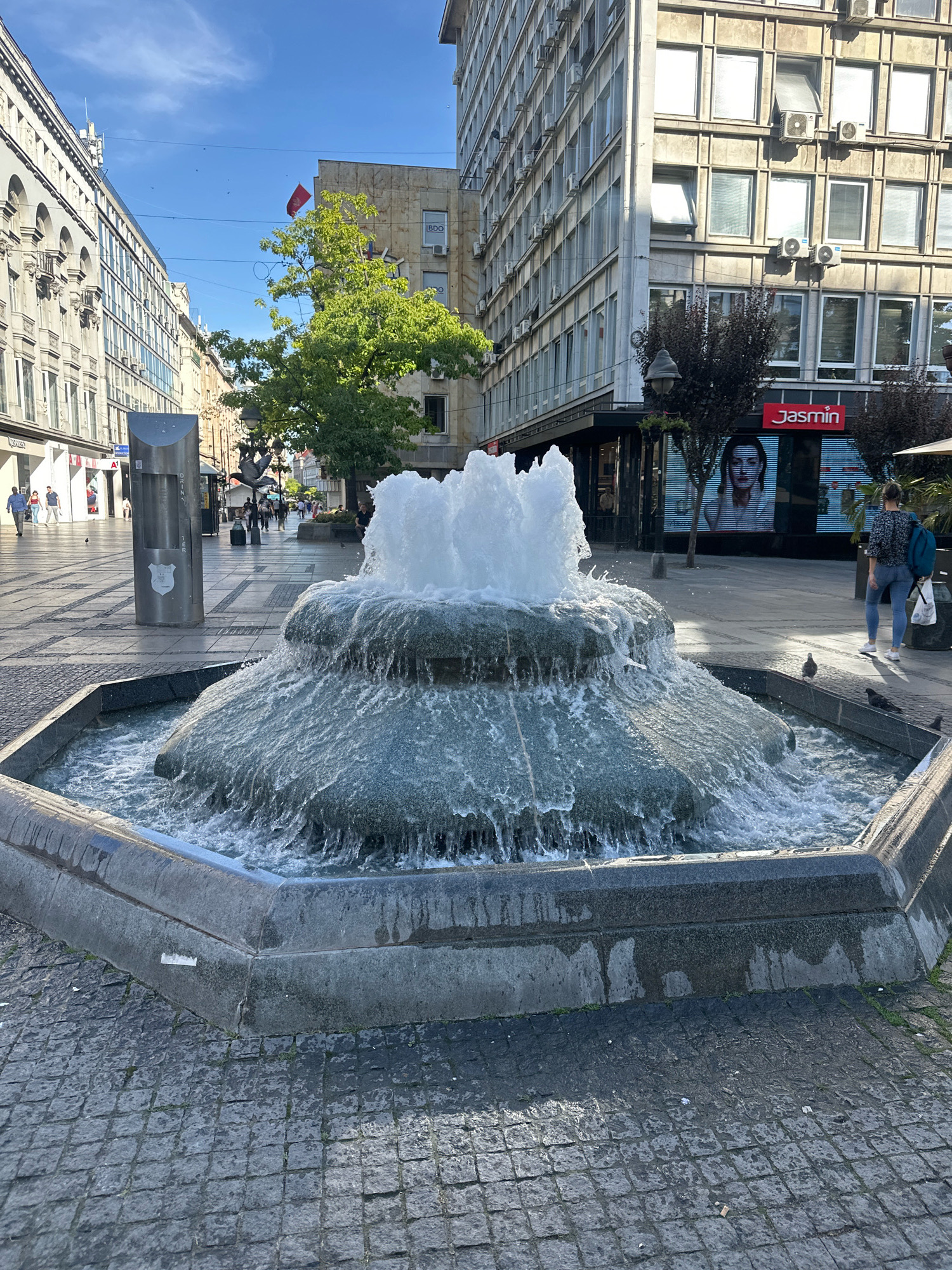 The image captures a street scene in Belgrade, Serbia, centered around a multi-tiered fountain with water cascading down its sides. The fountain is positioned on a cobblestone plaza with a hexagonal stone border. In the background, buildings line the street, one of which features an advertisement for "Jasmin" with a portrait of a woman. A person with a backpack is walking away from the fountain towards the right. Further down the street, other pedestrians are visible, and a red flag is flying from a building. The sky is blue with scattered clouds. The scene appears to be a public pedestrian area in an urban environment.