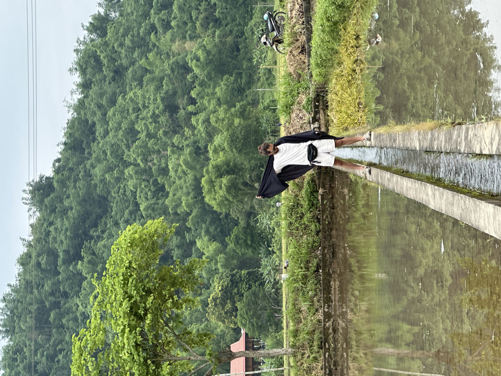 A young man with a beard and sunglasses stands on a narrow concrete path between two flooded rice paddies in Mai Chau, Vietnam. He wears a white t-shirt, white shorts, and a black fanny pack. He is holding a dark blue or black garment draped over his shoulders and his arms. In the background, lush green trees cover a hillside. A motorbike is parked on the right side of the scene. The sky is overcast. The water in the paddies reflects the surrounding greenery.