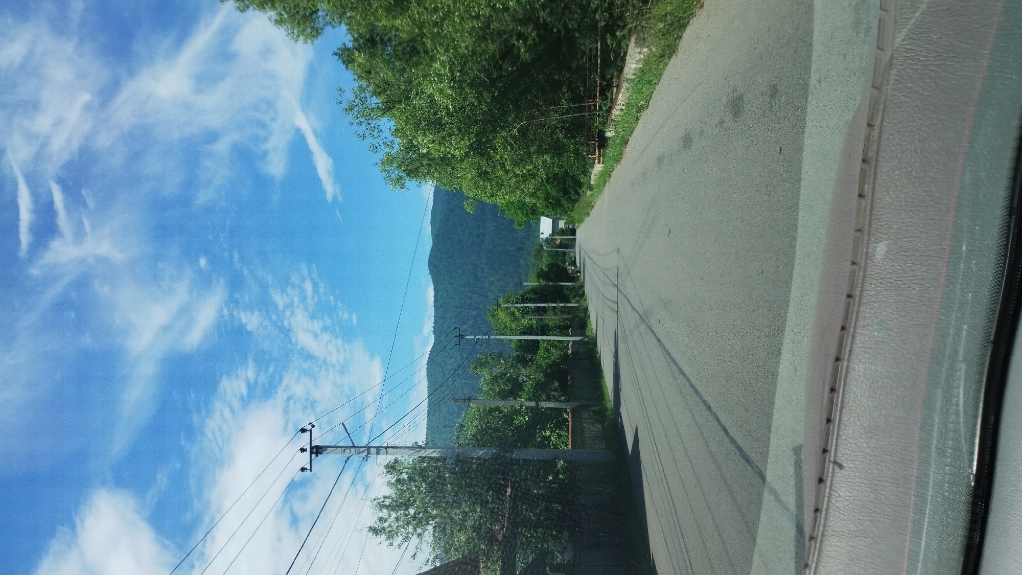 The image shows a view from inside a vehicle looking out at a paved road in Mizhhiria, Ukraine. The road curves to the right and leads towards a densely forested mountain range in the distance. Overhead, a bright blue sky is dotted with wispy white clouds. On either side of the road, there are trees and houses, and power lines run along poles. The foreground of the image shows the dashboard of the vehicle.
