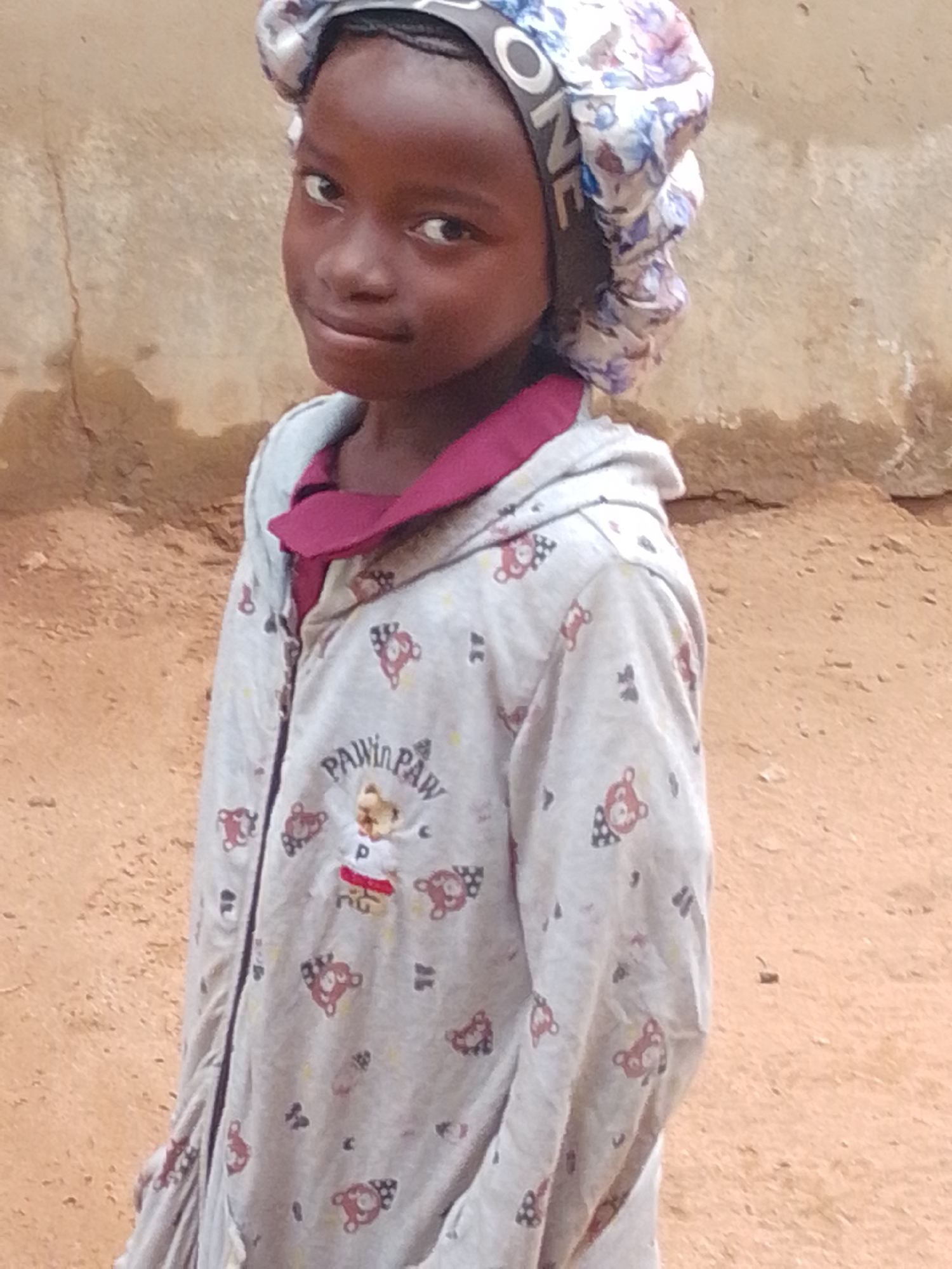 A young girl stands in front of a textured, light brown wall. She is looking directly at the camera with a slight smile. She has dark skin and dark hair, partially covered by a bonnet with a floral pattern and some text. She is wearing a light gray hooded jacket with a dark pink collar. The jacket is decorated with a repeating pattern of cartoon bears. The ground in front of her appears to be made of dirt or sand. The overall lighting suggests it is daytime. The location is unknown.