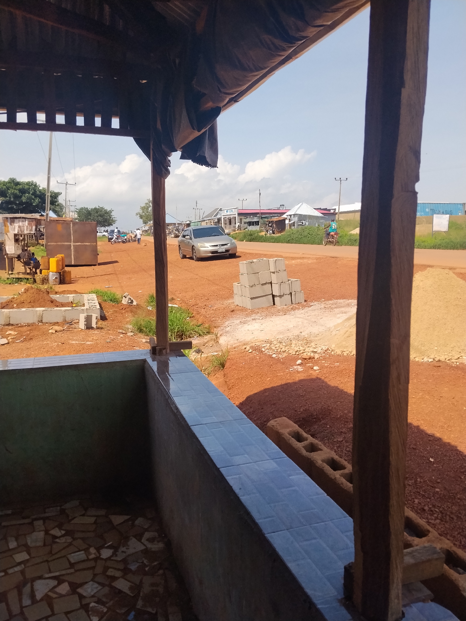 The image depicts a street scene in Durumi, Nigeria. In the foreground, a tiled ledge of an unfinished structure is visible, with debris scattered on the ground. Beyond this, a dirt road is present, with several people and a motorcycle. Construction materials, including concrete blocks and piles of dirt, are scattered on either side of the road. A silver sedan is parked on the right side of the road. In the background, there are several buildings, some appearing to be shops or small businesses, along with utility poles and trees. The sky is clear with some scattered clouds. The overall impression is of a developing area with ongoing construction and daily life unfolding.