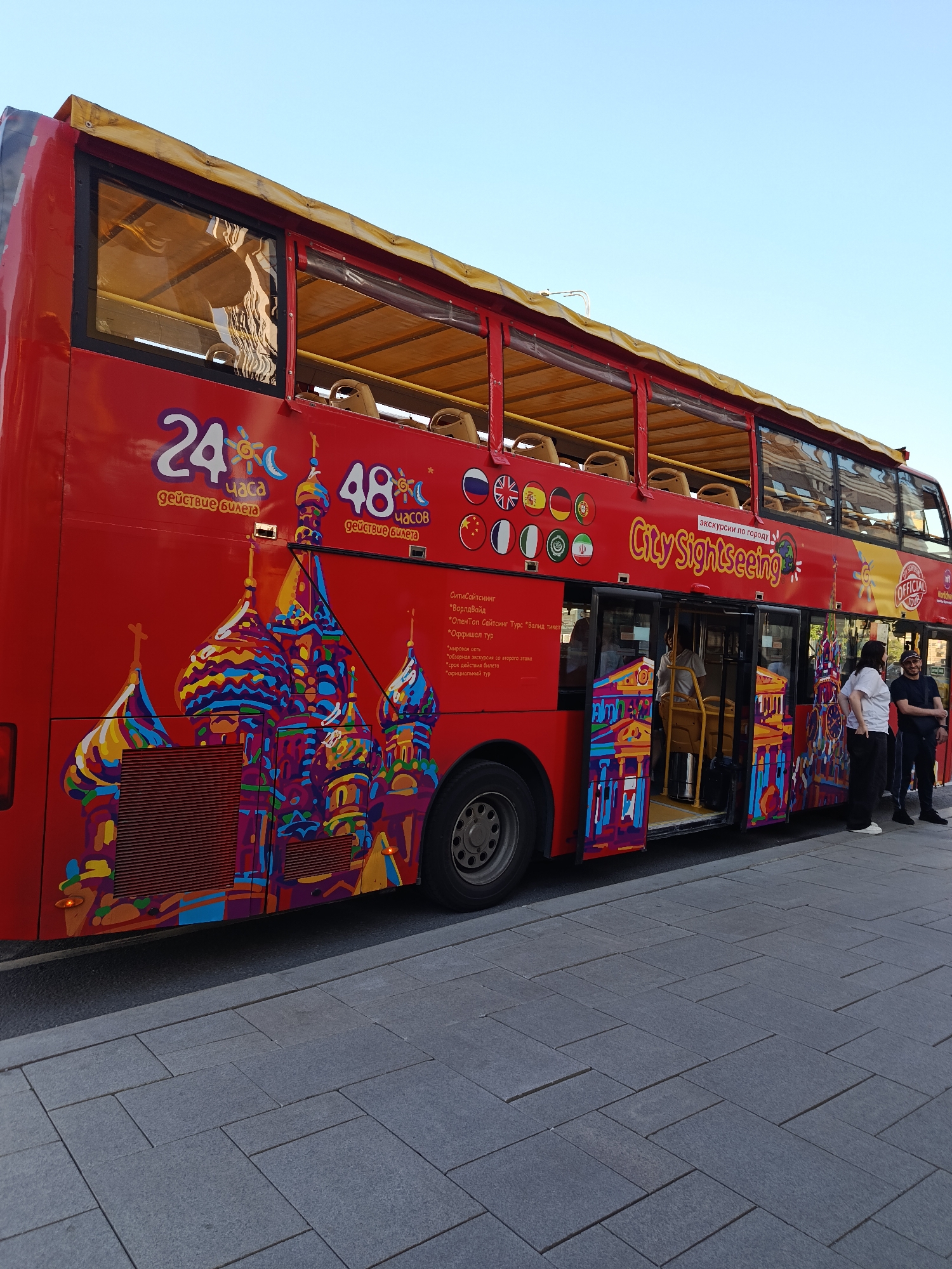 A red double-decker City Sightseeing tour bus is parked on a street next to a sidewalk paved with dark gray rectangular tiles under a clear blue sky. The bus features a vibrant, multi-colored graphic of a building resembling St. Basil's Cathedral. The side of the bus displays text in Russian indicating "24 hours ticket validity" and "48 hours ticket validity," along with "City Sightseeing" and "ЭКСКУРСИИ ПО ГОРОДУ" (City Tours). A row of national flags, including those of Russia, the UK, Spain, Germany, France, China, Italy, and Iran, are visible, suggesting multi-lingual services. The bus's front door is open, revealing a yellow interior pole and steps.
Three individuals are present near the open bus door. One person, wearing a light-colored top, is partially visible inside the bus. On the sidewalk, a woman with dark hair, wearing a white t-shirt and dark pants with white shoes, stands facing towards the bus. Beside her, a man with a beard and a dark baseball cap, dressed in a dark t-shirt and dark pants, stands with his arms crossed, also facing the bus. A third man, largely obscured by the second man, is partially visible behind him, also in dark clothing. The upper deck of the bus is open, with empty yellow seats visible and a yellow canvas canopy overhead. The environment is bright and sunny. The specific city and country are unknown.