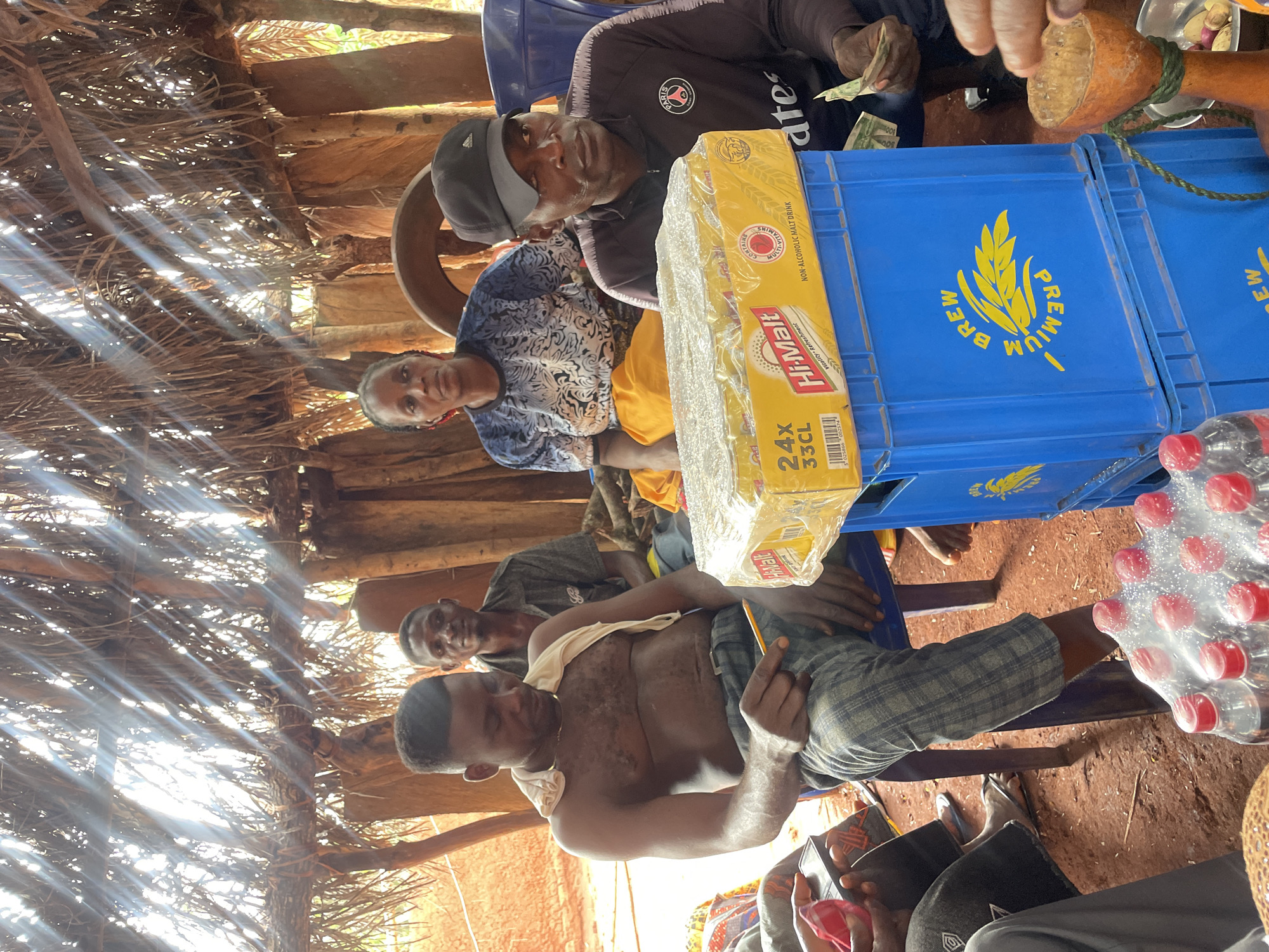 The image shows a gathering of people in what appears to be a rural setting in Ogbe, Nigeria. There are several individuals seated, including a shirtless man in the foreground with a cloth draped over his shoulder. A woman in a blue and white patterned shirt is also visible. There is a box of "Hi-Malt" malt drink on a blue crate labeled "PREMIUM BREW". Another crate is next to it, and there are plastic bottles of water nearby. Some individuals appear to be handling money. The location appears to be a hut or shelter with a thatched roof.