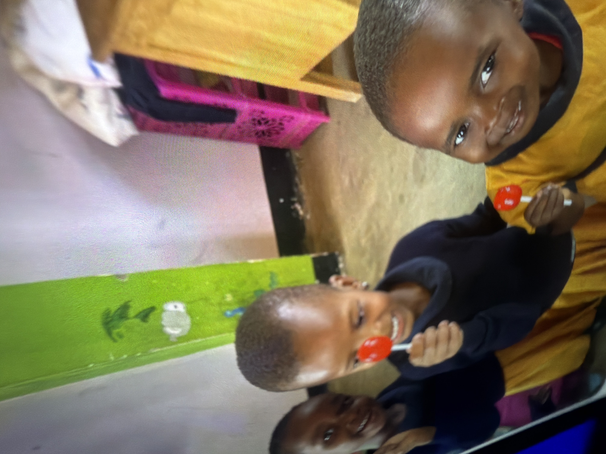 Three young children are in the foreground of a room. They are all smiling and holding red lollipops. The child in the center has a lollipop close to their mouth, while the child on the right is holding theirs out towards the viewer. The child on the left is mostly out of frame but appears to be smiling and reaching out. They are all wearing dark-colored tops and yellow skirts or pants. The room has a green wall with some drawings on it, and a wooden desk or table in the background. The floor is a light brown color.