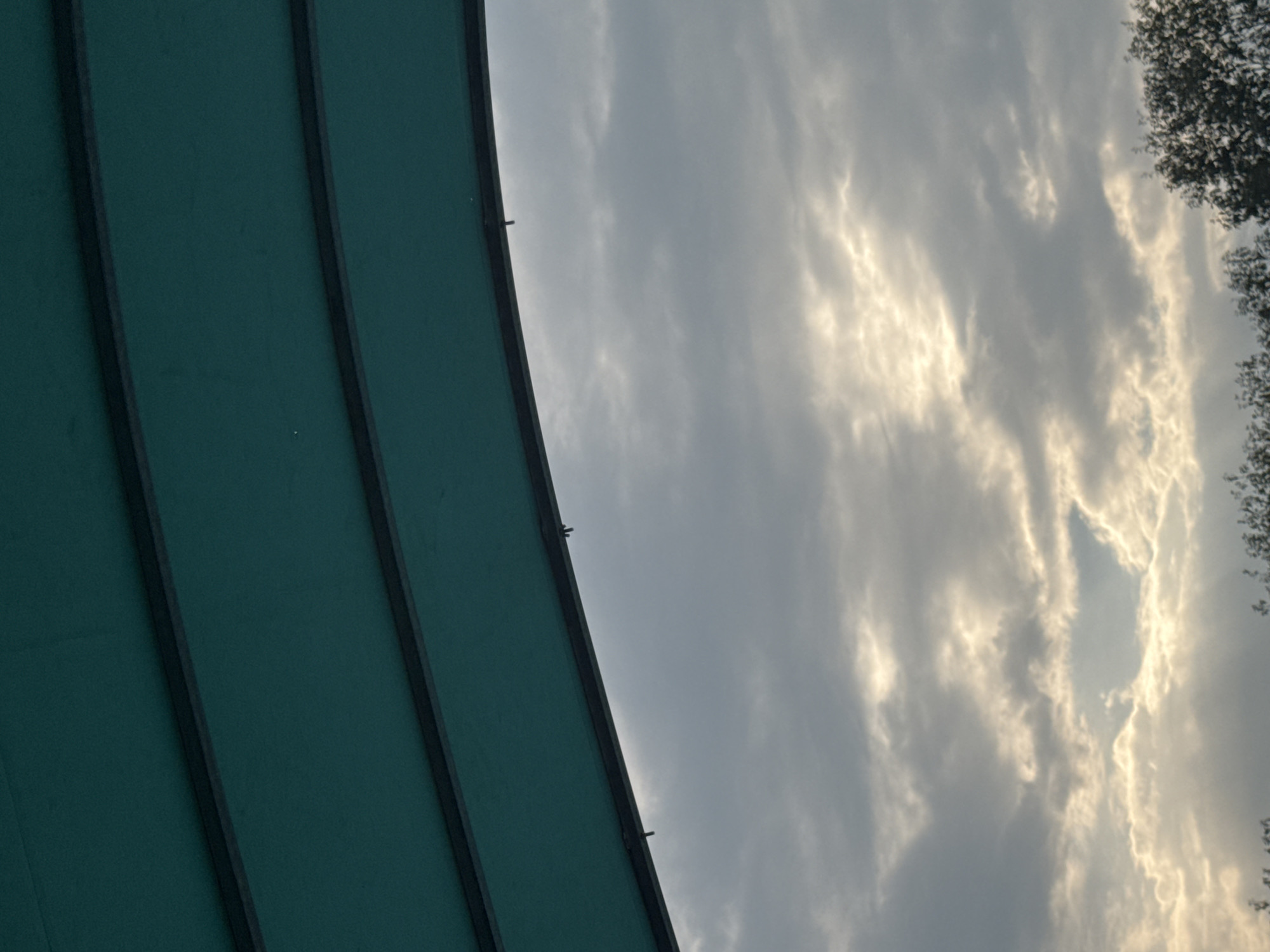 The image displays a cloudy sky viewed from beneath a teal-colored awning supported by dark frames. The awning occupies the top portion of the frame, angled downwards from left to right. The sky is filled with scattered clouds, with areas of brighter light indicating sunlight filtering through. The bottom edge of the image shows a blurred line of dark green foliage, suggesting trees or bushes. No people or specific landmarks of Chiasso, Switzerland, are visible.