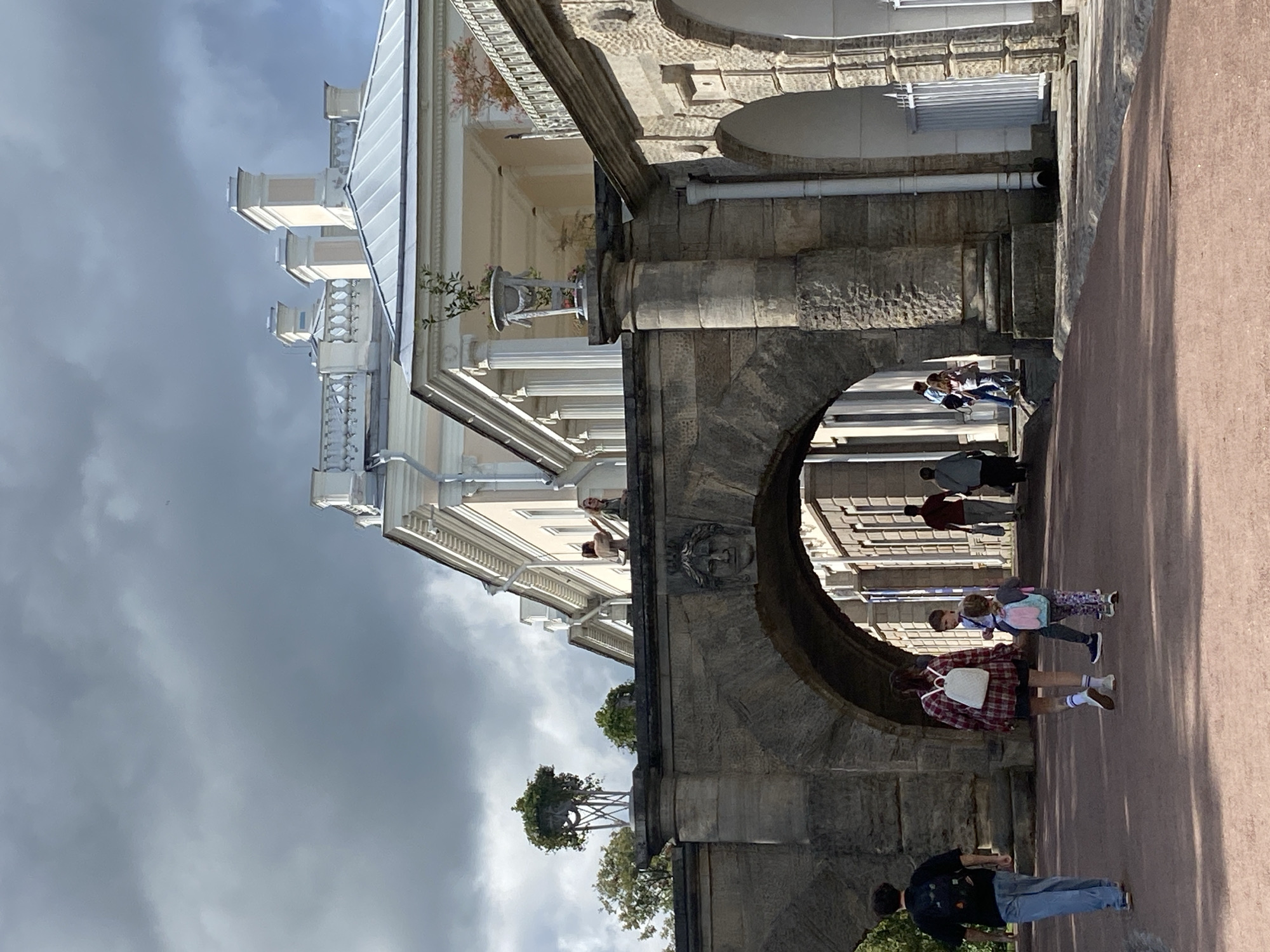 The image depicts an exterior view of the Catherine Palace in the city of Pudomyagi, Russia, under a partly cloudy sky. In the foreground, several individuals are walking on a reddish-brown path. A person in a black t-shirt and blue jeans walks away from the camera. Behind them, a person in a red and black plaid shirt and white shorts with a white backpack walks towards an arched stone structure. To their right, a child in a patterned outfit with a light blue backpack walks in the same direction. Further back, several other individuals are visible walking through the arched structure and beyond. The stone archway is adorned with a carved face. Beyond the archway, a grand building with white columns and ornate architecture is visible. A potted plant sits on a pedestal atop the stone structure. The overall scene captures a moment of pedestrian activity in the vicinity of a historical landmark.