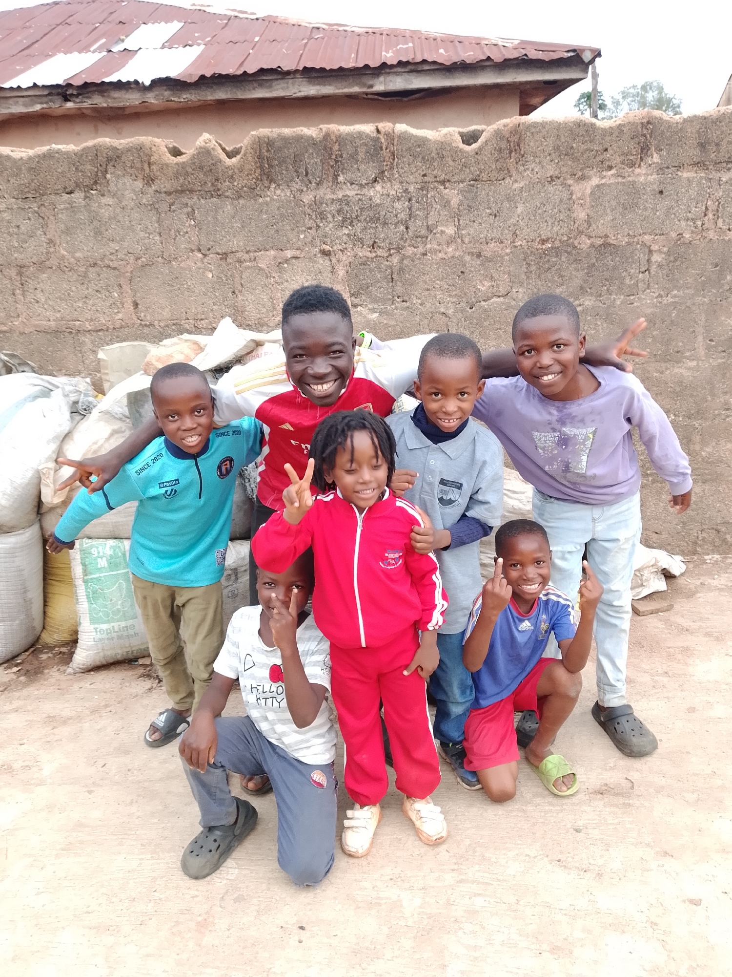 Eight children, appearing to be between the ages of five and twelve, pose for a photo in front of a textured, rough-hewn brick wall. The wall is topped with a corrugated metal roof. Behind the children, several large, white and tan sacks are piled up. The ground is a light brown, dusty earth. The children are dressed in casual clothing. Two of the children are making peace signs with their fingers. The overall impression is a candid snapshot of children in an outdoor, possibly rural or informal urban setting.