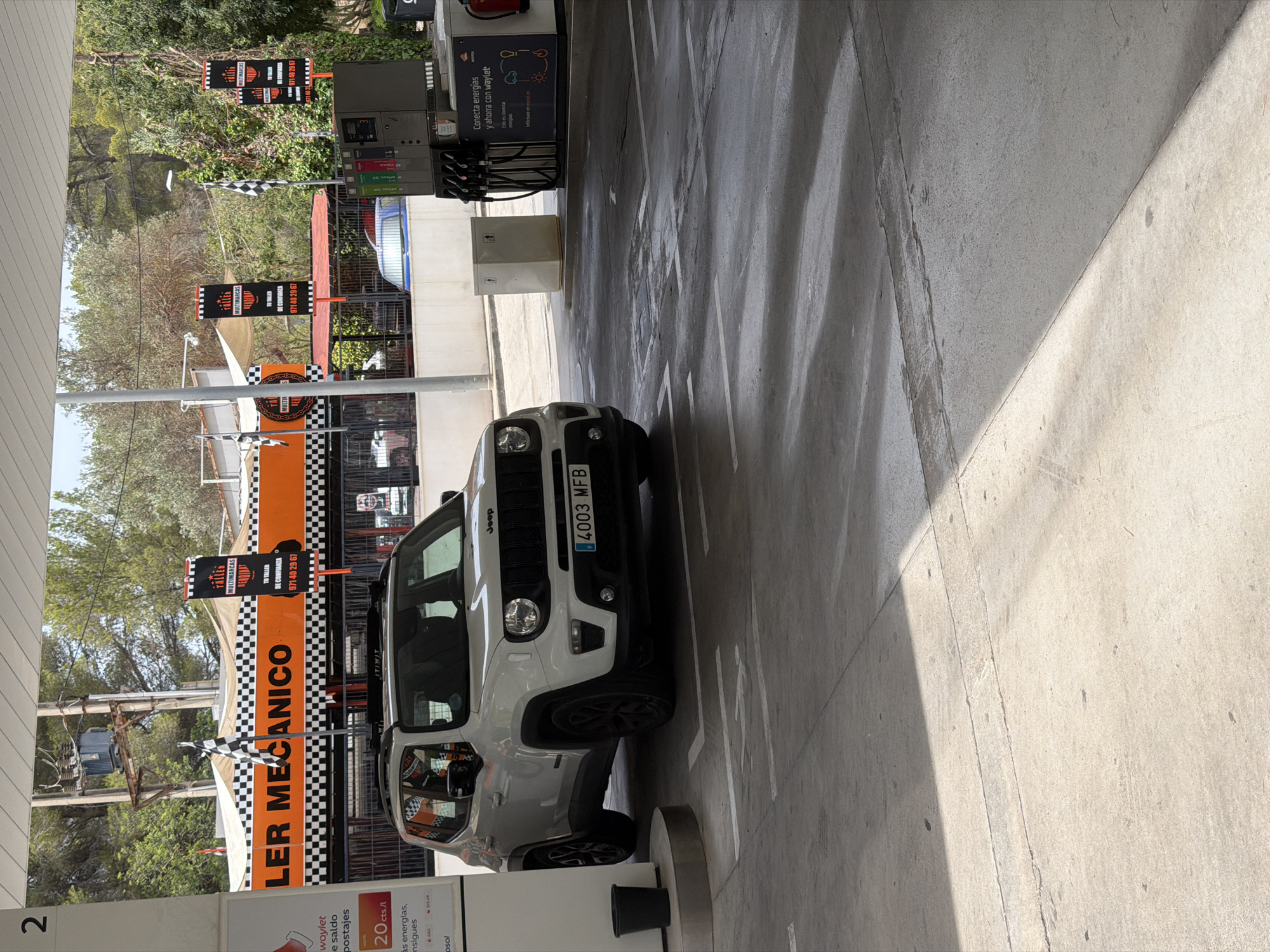 The image depicts a white Jeep Renegade with license plate 4003 MFB parked under the canopy of a gas station in Calvià, Spain. The gas station features an orange and black checkered facade with the words "TALLER MECANICO" and "TALLER MULTIMARCAS" visible on banners and signs. Racing flags are also present. To the right of the Jeep, a fuel pump station is visible, with signage indicating a promotion called "Conecta energias y ahorra con waylet." The ground is paved concrete, with white painted lines indicating parking or driving lanes. Trees and vegetation are visible in the background, suggesting an outdoor, possibly sunny environment. There are no people clearly visible in the image.