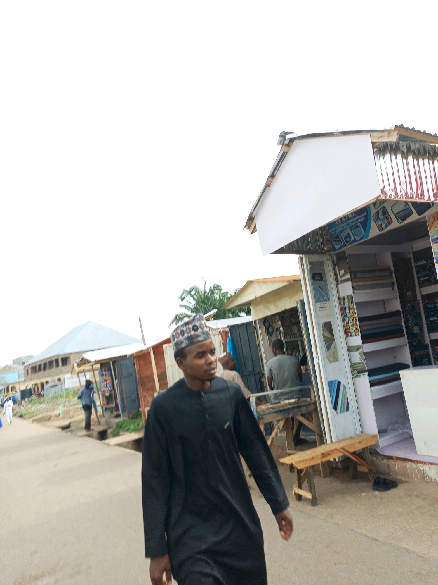 In Arawa, Nigeria, a busy marketplace scene unfolds. A man in a black traditional outfit and a patterned cap walks down a paved street, his gaze directed forward. Behind him, a row of open-air shops line the street, displaying textiles and various goods. In one shop, rolls of fabric are neatly arranged on shelves. Another man, wearing a grey t-shirt and dark pants, is visible inside a shop, possibly attending to customers. The environment is a typical urban setting with buildings and a clear, overcast sky. The street appears to be a thoroughfare for both pedestrians and possibly vehicles, though none are prominently featured in this particular shot.