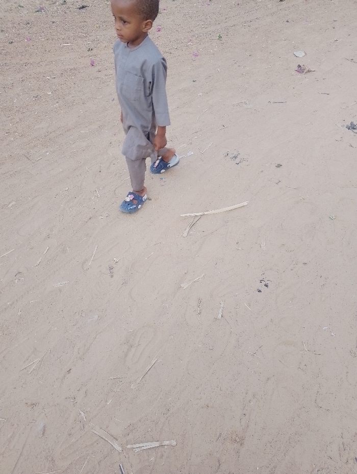 A young boy is walking on a sandy surface. He wears a gray outfit and blue sandals with floral decorations. The ground is covered with sparse pieces of debris, possibly dried twigs or leaves. He is walking to the left of the frame, looking in the same direction. The background appears to be an open, dry area.