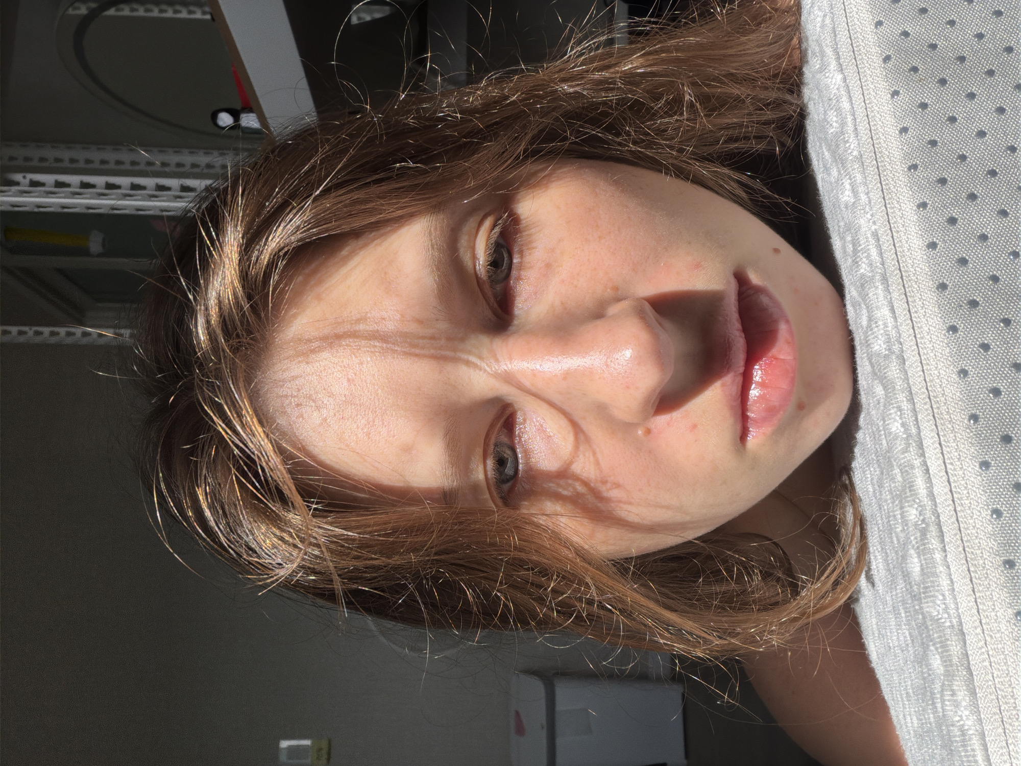 A close-up, eye-level, forward-facing shot of a young Caucasian female with light brown hair, freckles, and a pale complexion. She is looking directly at the camera with a neutral expression. Sunlight illuminates her face from the left, casting shadows across her forehead and nose. Her hair is slightly messy and appears to have some highlights from the sun. The background is dark, with hints of shelving units and a white bin. The lower part of the image shows her face resting on a textured, light-colored surface, possibly a mattress or cushion, with small, dark circular patterns.