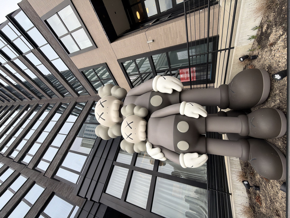 Two large-scale KAWS Companion sculptures are installed outdoors in front of a modern residential building in Long Island City, Queens, New York, United States of America. The sculptures depict two humanoid figures, one larger and one smaller, in shades of dark grey and light grey. The larger figure's left arm is around the shoulder of the smaller figure. Both figures have light grey heads with black "X" marks for eyes and mouth, white gloves, and two circular grey buttons on their lower torsos. They stand on a sparse landscaped bed containing soil, minimal dry vegetation, and several black ground-level spotlights.
The background consists of a multi-story building constructed from dark and light brown brick, featuring numerous large, rectangular windows with dark frames. The windows reflect the overcast sky and elements of the surrounding urban environment. A red "FOR LEASE" sign, displaying "RL" and the phone number (917) 302-2600, is affixed to a ground-level window on the lighter brick section of the building to the right of the sculptures. A black metal railing with horizontal bars is situated in front of this section of the building. The image is captured from a low-angle perspective, emphasizing the height of the building behind the sculptures.