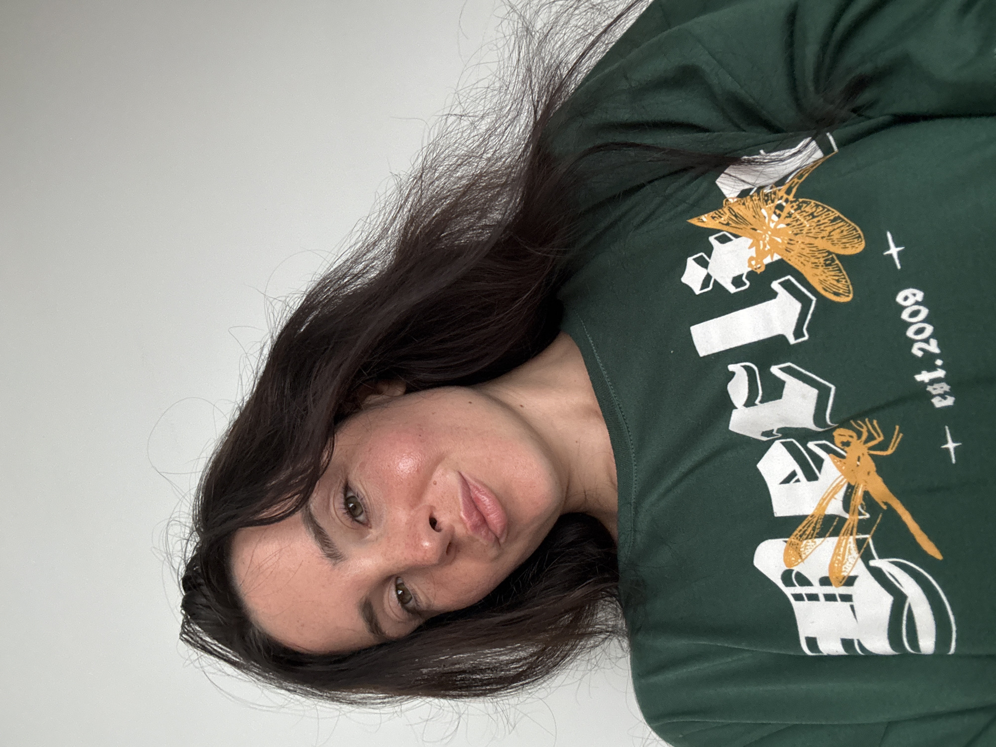 A selfie of a woman with dark hair and light skin. She is wearing a dark green long-sleeved shirt with a graphic design of white lettering and two orange dragonflies. Her hair is dark brown and long, with some strands falling across her face. She is looking at the camera with her head tilted slightly and her lips pursed. The background is a plain white wall. The scene is indoors, likely in a residential setting in North Kuta, Indonesia.