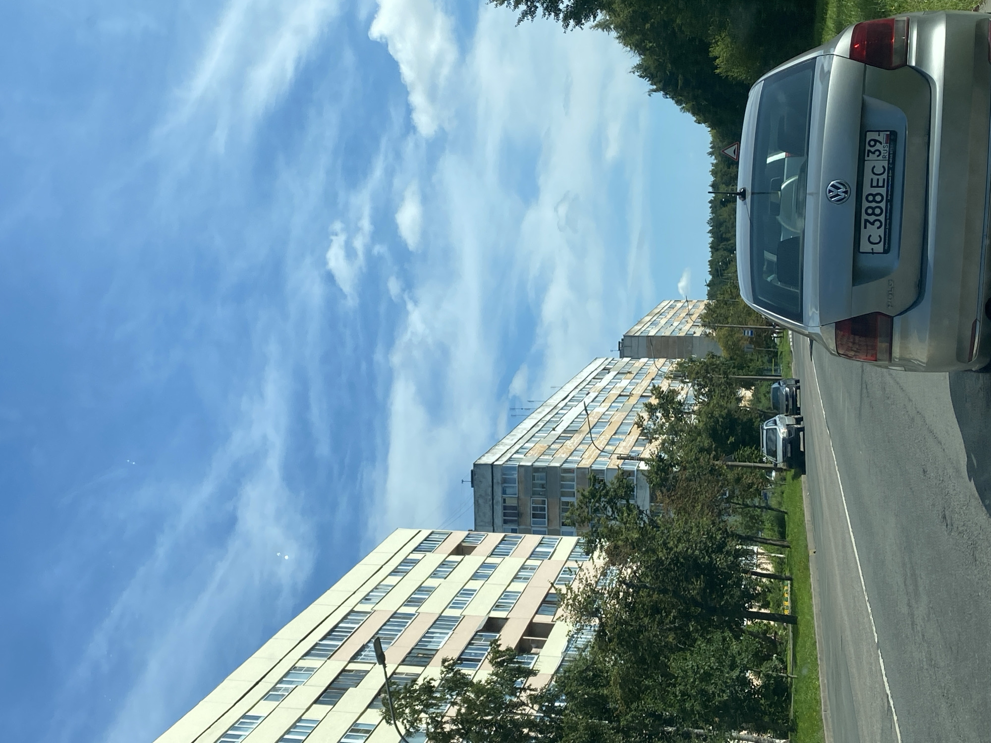 The image shows a street in Lomonosov, Russia, with residential buildings lining the left side and a dense forest on the right. In the foreground, a silver Volkswagen Polo sedan is visible from the rear, its license plate reading "C 388 EC 39 RUS". The car is on an asphalt road with a white line marking the lane. Several other cars are seen further down the road, including a silver SUV and a dark-colored sedan. Trees and streetlights are present along the sidewalk. The sky is blue with scattered white clouds.