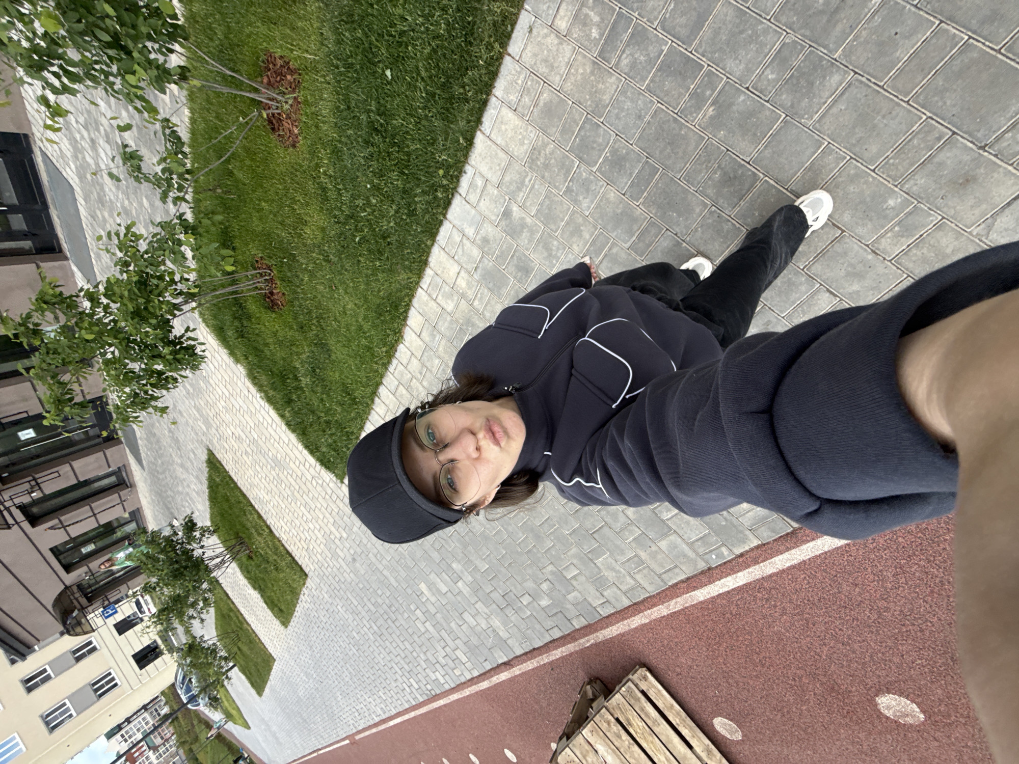 A selfie of a person wearing glasses and a black cap and jacket with white lines, standing on a gray brick walkway bordered by grass and trees, with a red running track nearby and buildings in the background. The location is likely Pervomaysky, Russia.