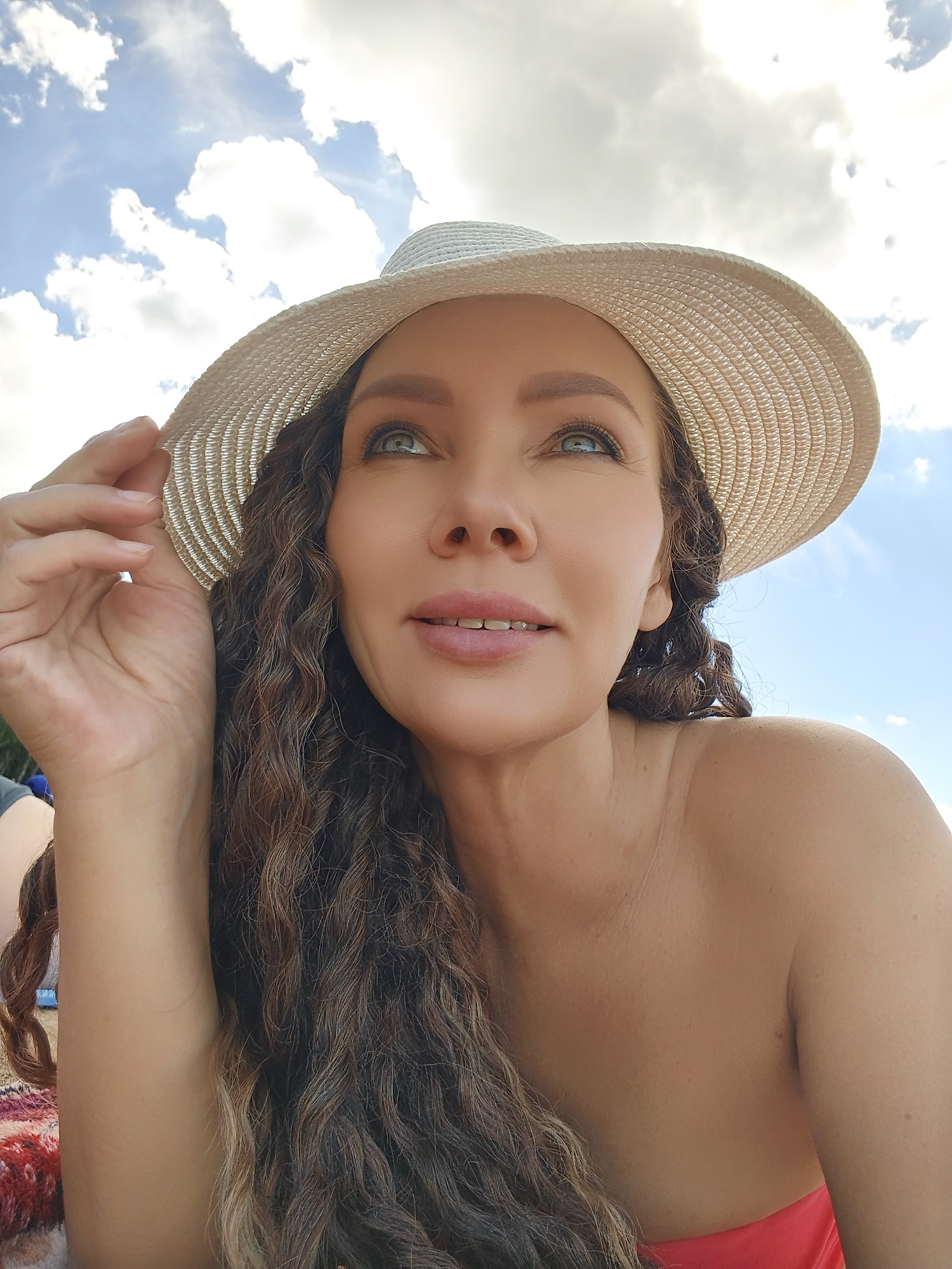 A woman with long, curly brown hair and blue eyes, wearing a white straw hat and a red top, looks up at the sky. The woman is holding the brim of her hat with her right hand. The sky is blue with white clouds and the sun is shining. The image appears to be taken outdoors, possibly at a beach or park, given the natural light and the casual attire. The woman's expression is serene and relaxed, suggesting a pleasant day. There is no visible text in the image.