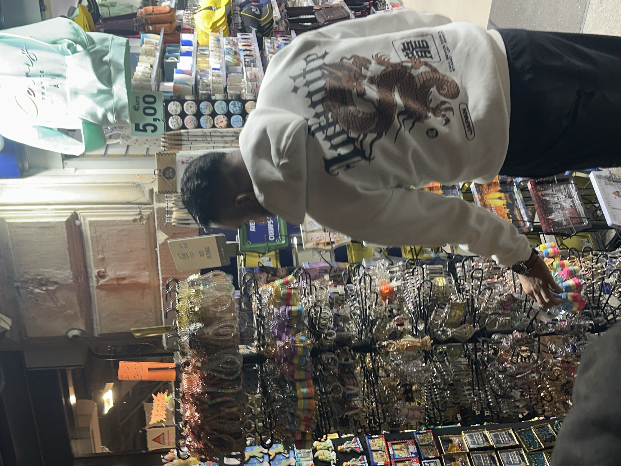 An individual is captured in a souvenir stall, likely in Paris, France, browsing various items. The image appears to be taken under artificial light, suggesting it could be an evening scene or an indoor market setting.
The main subject, a male figure, is seen from behind and slightly to the side, wearing a white long-sleeved shirt adorned with a brown dragon design and Japanese characters (龍, meaning 'dragon'). The word "JAPAN" is also visible on his sleeve. He is dressed in black pants and wears a watch on his left wrist. He is bent over, actively reaching his right hand towards a display of numerous colorful items, indicating he is selecting or examining merchandise.
The stall is densely packed with a wide array of souvenirs. Prominently displayed are multiple racks filled with various keychains, charms, or small ornaments, many clear plastic or vibrantly colored. Below these are rows of small, square items, possibly magnets or postcards. In the upper left, a light blue hoodie or sweatshirt hangs, bearing the text "FRANCE" and "Cœurs," with an attached price tag clearly showing "Euro 5,00." Other items visible include bags or backpacks in green, yellow, and brown tones, and shelves stocked with packaged goods.
Location context is hinted by the "FRANCE" text and the "Euro 5,00" price tag. The OCR also identified "CHAMPS," which likely refers to the Champs-Élysées, a famous avenue in Paris. The atmosphere suggests a typical souvenir shopping experience.