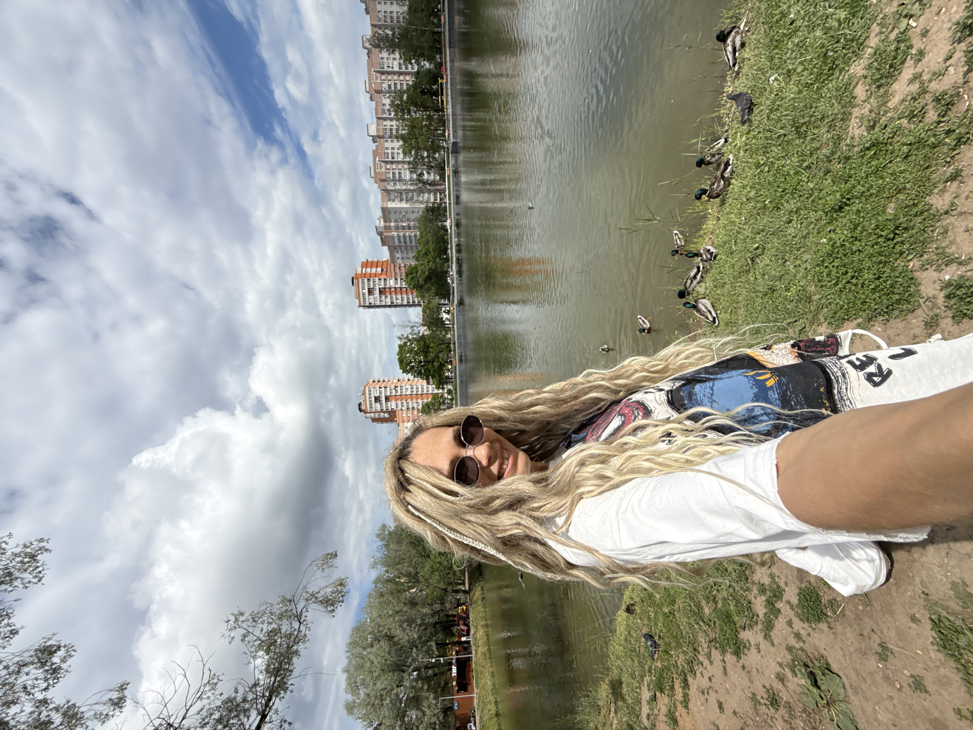 A selfie captured in a bright, daytime setting shows a person with long, blonde dreadlocks and sunglasses smiling at the camera. They are wearing a white t-shirt with a graphic design, part of which shows the letters "RE". The person's arm, holding the camera, is visible in the foreground.
The background features a tranquil lake or pond with several ducks resting on the grassy bank and swimming in the water. On the opposite side of the water, a line of multi-story apartment buildings with light-colored facades and reddish-brown sections is visible, suggesting an urban or suburban park setting. Trees line the far bank, and a cloudy but bright sky stretches above the buildings. The overall impression is one of a pleasant day spent outdoors by the water.