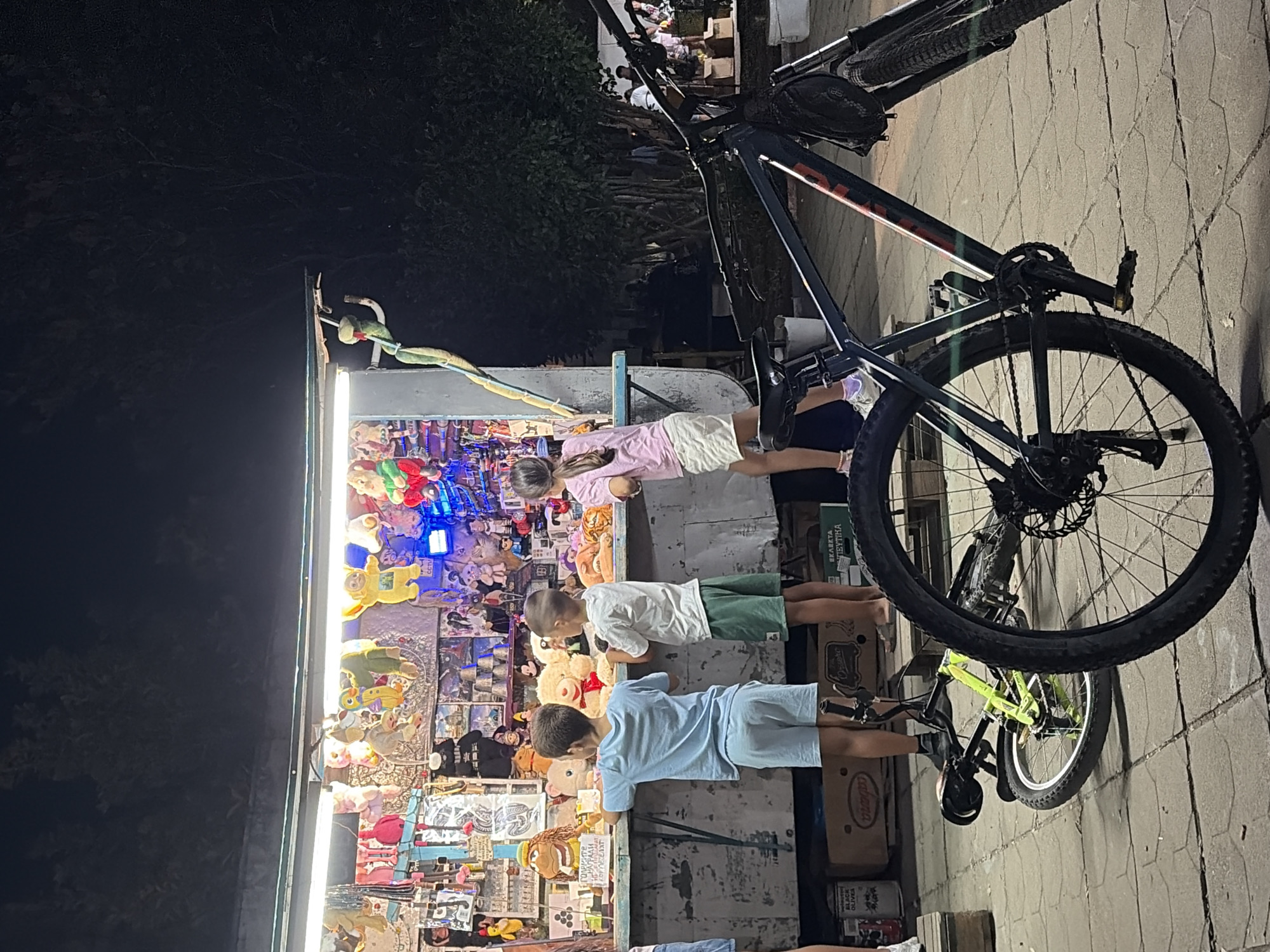 At night in Rupite, Bulgaria, three children stand at a brightly lit souvenir stand, examining plush toys. A boy in a blue t-shirt and shorts stands on the left, a boy in a white t-shirt and green shorts in the middle, and a girl in a pink t-shirt and white shorts on the right. The stand displays a variety of stuffed animals and other trinkets. In the foreground, two bicycles are parked, one upright and dark-colored, the other leaning and neon green. The ground is paved with square tiles, and trees are visible in the background.
