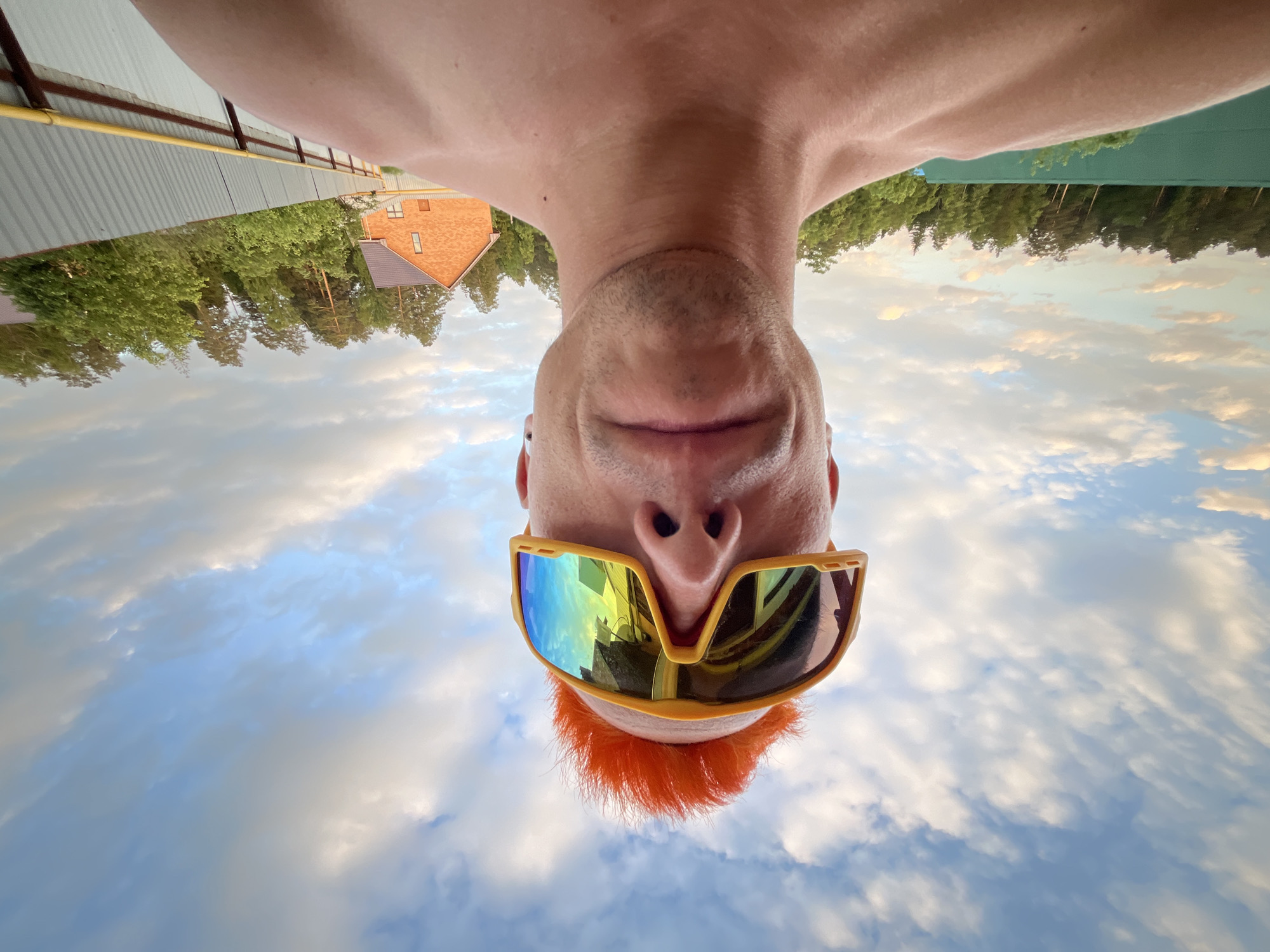 A man with orange hair and sunglasses is pictured from a low-angle perspective against a backdrop of sky and clouds. Upside-down, the image shows a building and trees in the background. The photograph was taken in Chaikovsky, Russia.