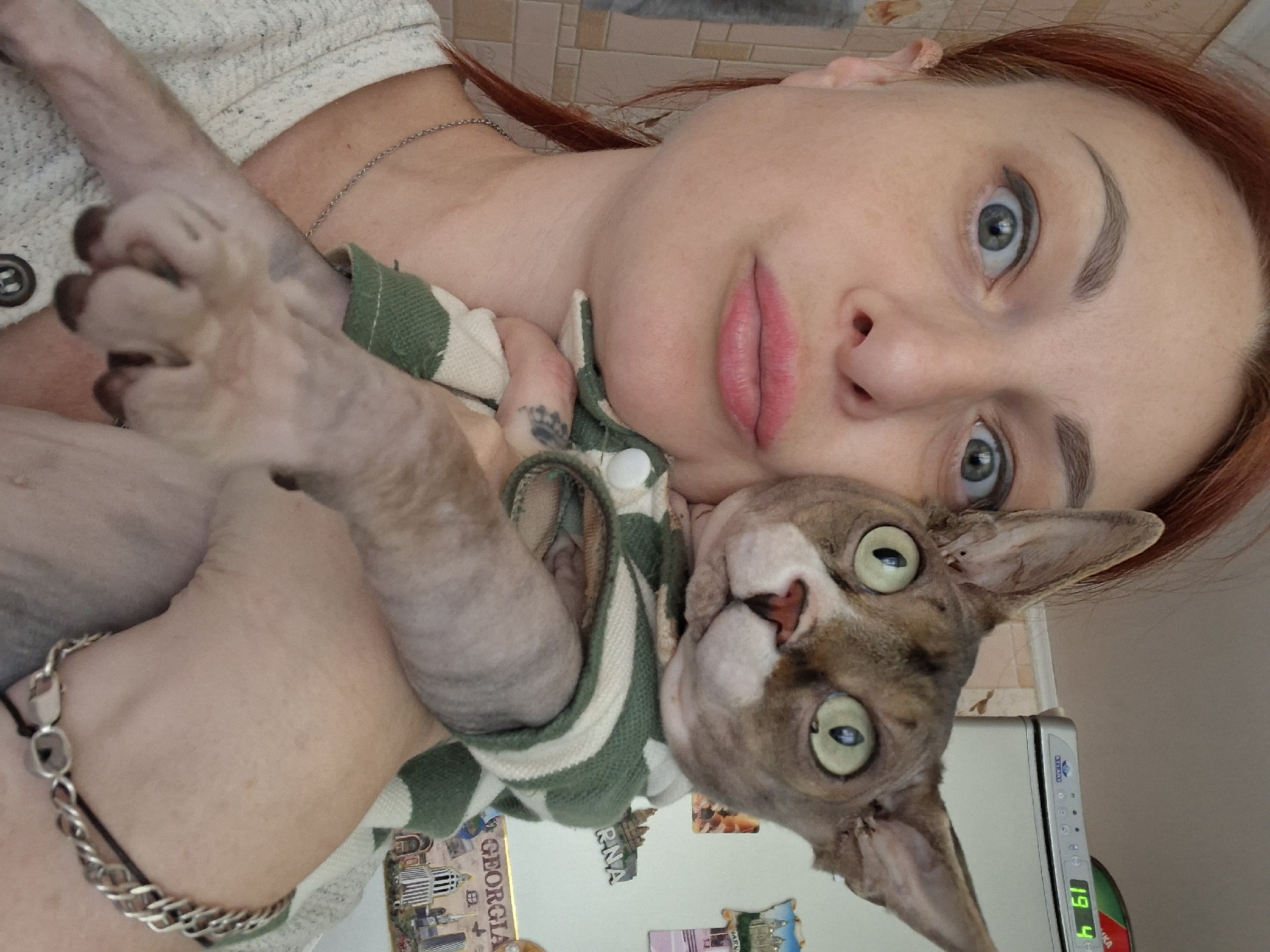 A woman with reddish hair and blue eyes is taking a selfie with a hairless cat. The cat is wearing a green and white striped shirt. They are in a kitchen, with a refrigerator visible in the background. The refrigerator has magnets on it, including one that says "GEORGIA". The woman is wearing a light-colored sweater and a silver necklace. The cat has large, green eyes and pointed ears. The woman is making a pouty face. The scene is indoors and appears to be a casual moment. The location of Myadzyel, Belarus is not explicitly depicted or suggested by any visible elements in the image.