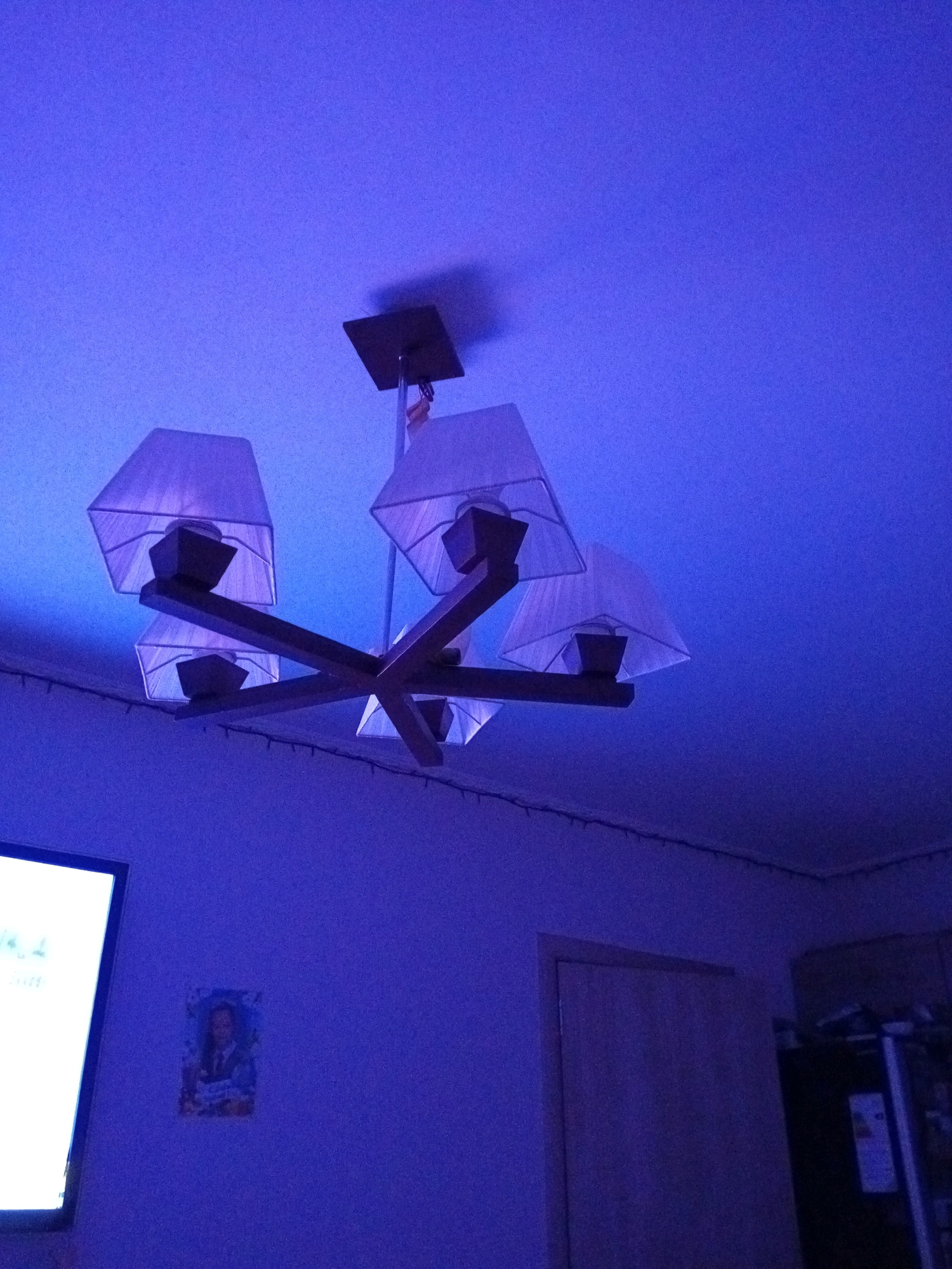 An indoor shot shows a chandelier hanging from a blue ceiling. The chandelier has a wooden base and six fabric lampshades. The lighting is predominantly blue, creating a dim and atmospheric environment. In the lower left corner, a portion of a bright white television screen is visible. On the wall to the right of the TV, a framed picture of a person is hanging. Further to the right, a closed wooden door is visible, and beyond that, the edge of a dark appliance, possibly a refrigerator, can be seen. The overall impression is that of a room with low ambient lighting, possibly a living room or entertainment area. The specific location of Petropavlovsk-Kamchatsky, Russia, is not directly evident from the visual information presented in the image.