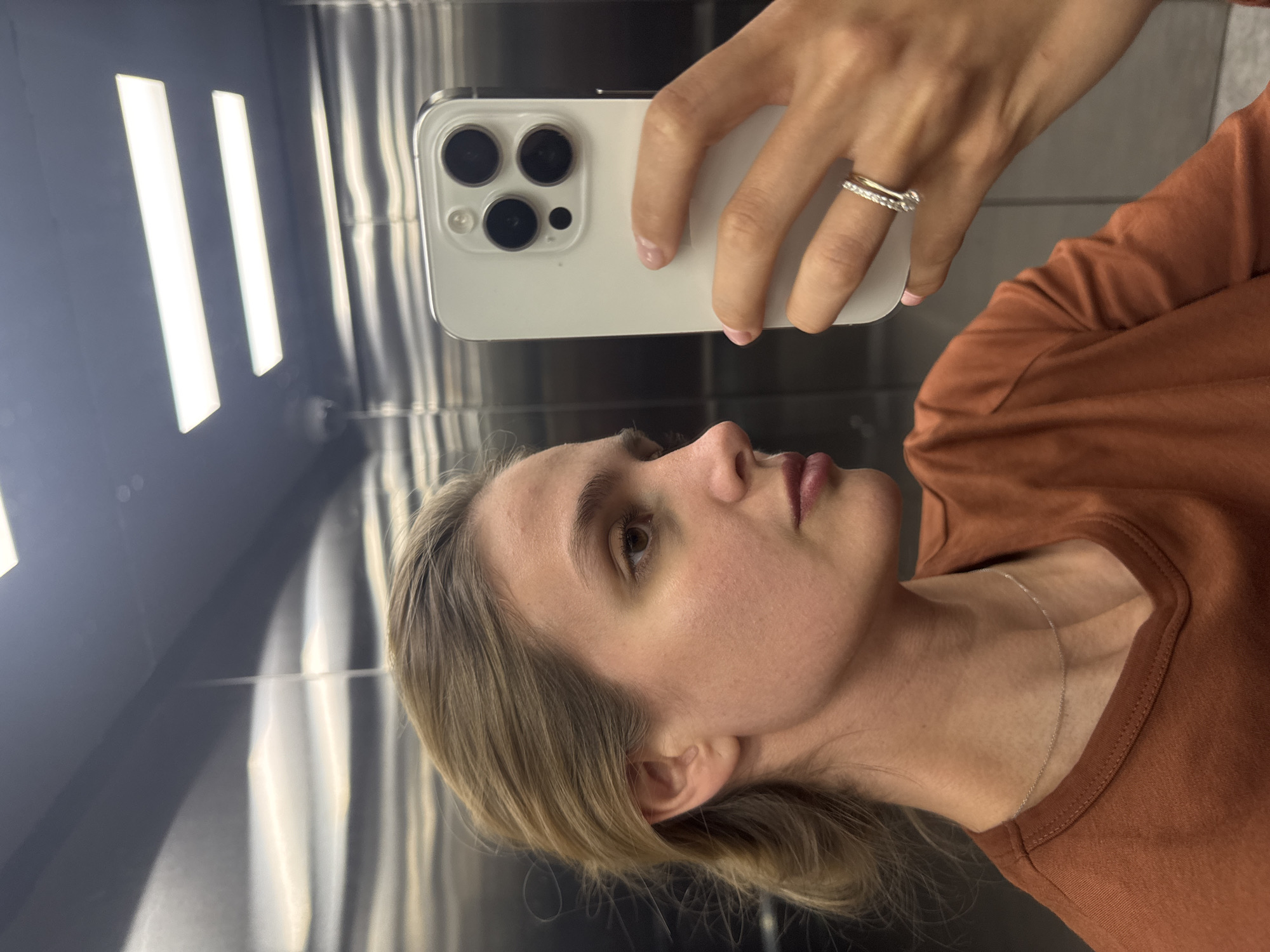 A woman with blond hair takes a selfie in an elevator. She is wearing a copper-colored shirt and a thin silver necklace. Her phone is white and has three camera lenses. The elevator has a stainless steel interior with vertical light fixtures. The location is Unknown, Unknown.