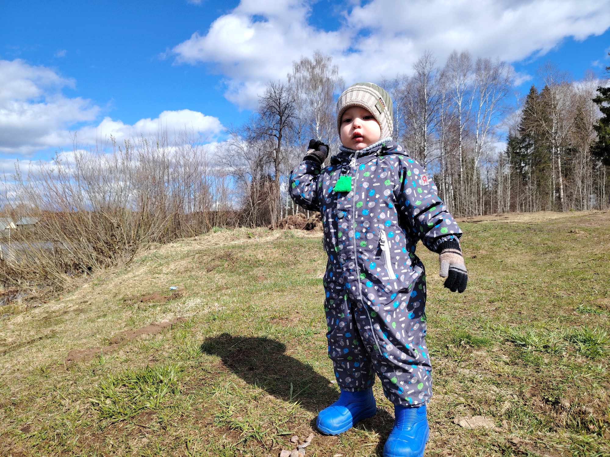 A toddler stands in a grassy field, wearing a patterned grey snowsuit, blue boots, and a knitted hat with matching mittens. The child has one hand raised to their face, and the other is at their side. Behind the child is a backdrop of trees and a blue sky with scattered clouds. In the background is a grassy hillside and some bare trees. The scene appears to be outdoors during daylight. The location seems to be a rural or park area in the city of Unknown, Unknown.
