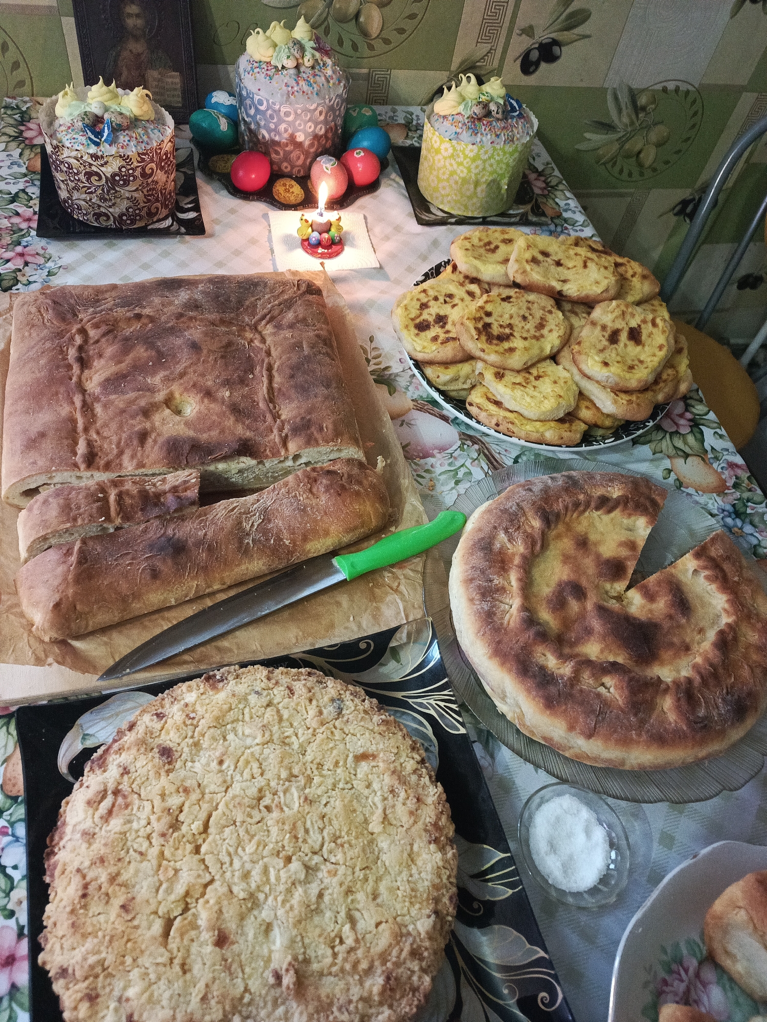 The image depicts a table laden with food, likely for a celebration, possibly Easter, given the presence of decorated Easter cakes (kulich) and colored eggs. The table is covered with a floral patterned tablecloth. Located at the center of the table is a square flatbread cut with a knife, next to a plate of cheese pies, and a round flatbread with a slice cut out. At the edge of the table is a cake with a crumbly topping. A small dish of white powder is on the table, as well. In the background, there are two more Easter cakes and colored eggs around a small candle. There is a religious icon next to the cakes. The location of this image is believed to be Chaikovsky, Russia. No people are visible in the image. The setting appears to be indoors, possibly a home.