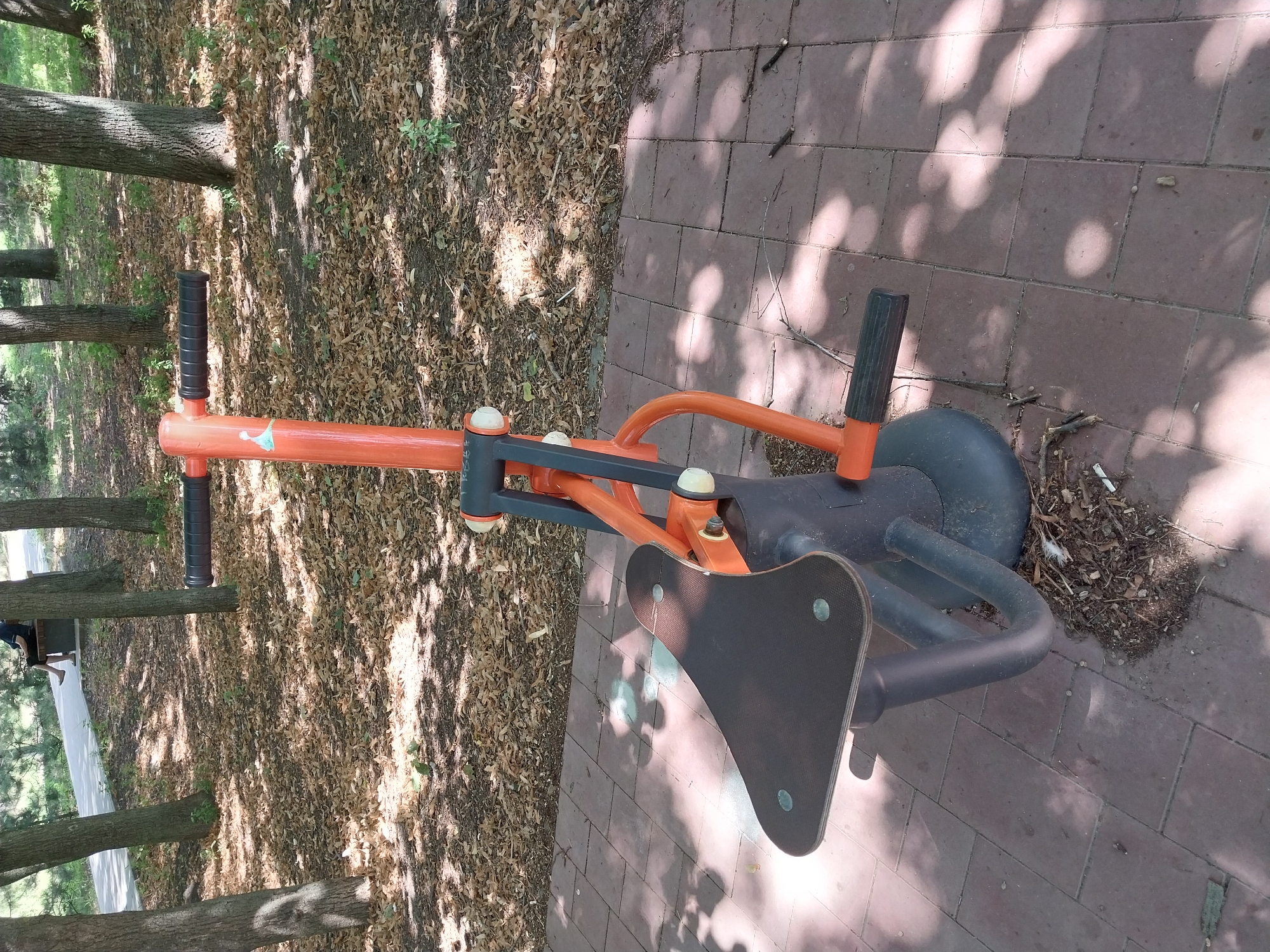 The image shows an outdoor fitness machine in a park setting. The machine, which appears to be a leg press or similar equipment, is predominantly orange and black with a dark gray, bulbous base. It is situated on a paved area made of reddish-brown rectangular tiles, with a scattering of leaves and small branches on the ground around the base of the machine. In the background, there are trees with dappled sunlight filtering through their leaves, casting shadows on the ground. Further in the distance, a person is seated on a bench on a paved walkway. The environment suggests a public park, likely in the city of Dnipro, Ukraine, given the context.