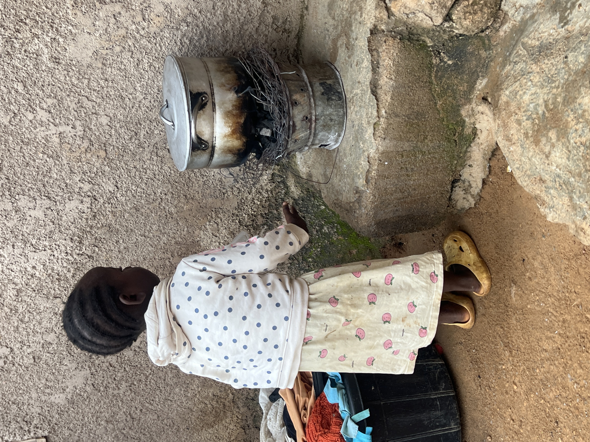 A young girl, seen from the back and right side, stands by a rustic cooking setup. She has dark skin and her hair is styled in cornrows. She is wearing a white hoodie with blue polka dots and a light-colored skirt adorned with pink strawberry-like patterns. On her feet, she wears yellow slip-on shoes.
Her left hand is extended towards a metal pot that sits atop a charcoal stove. The stove is made of stacked metal cylinders, with visible burning charcoal inside. The pot is stained from cooking. To the left of the girl, a black plastic container is filled with various items of clothing, including an orange knitted item and a blue cloth.
The scene appears to be outdoors, with a textured, concrete-like wall behind the girl and the cooking setup. The ground is a reddish-brown dirt. To the right of the stove, there is a rough, stone-like surface. The overall environment suggests a utilitarian, possibly residential, outdoor space.
While the image depicts a scene that could be found in Jos, Nigeria, the specific location within the city cannot be determined from the visual information alone.