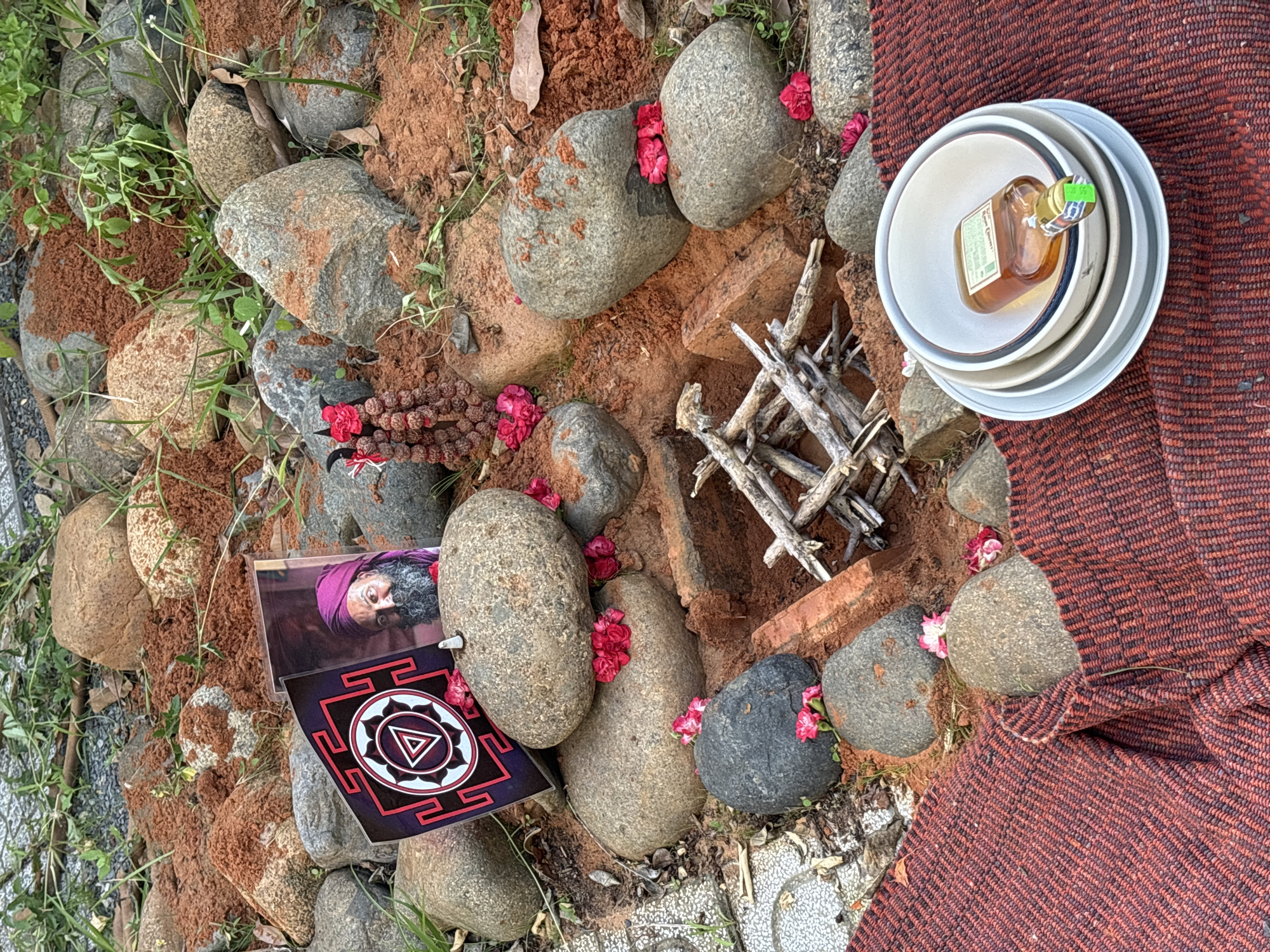 A close-up, outdoor, eye-level shot shows a collection of ritualistic items arranged on the ground, surrounded by rocks and red soil. In the center, a small pile of dried twigs forms a pyre-like structure within a shallow pit. To the left of the pyre, an open book displays a geometric symbol and a portrait of a man with a purple turban and beard. Nearby, a string of dark brown beads, possibly rudraksha, hangs from a red flower. Scattered around the rocks are small pink flowers. In the foreground, a stack of white and gray bowls is placed on a red and black patterned cloth. A small, amber-colored bottle with a green label rests inside the top bowl. The environment appears to be a natural setting with rocks of varying sizes, some covered in red soil, and patches of green grass.
