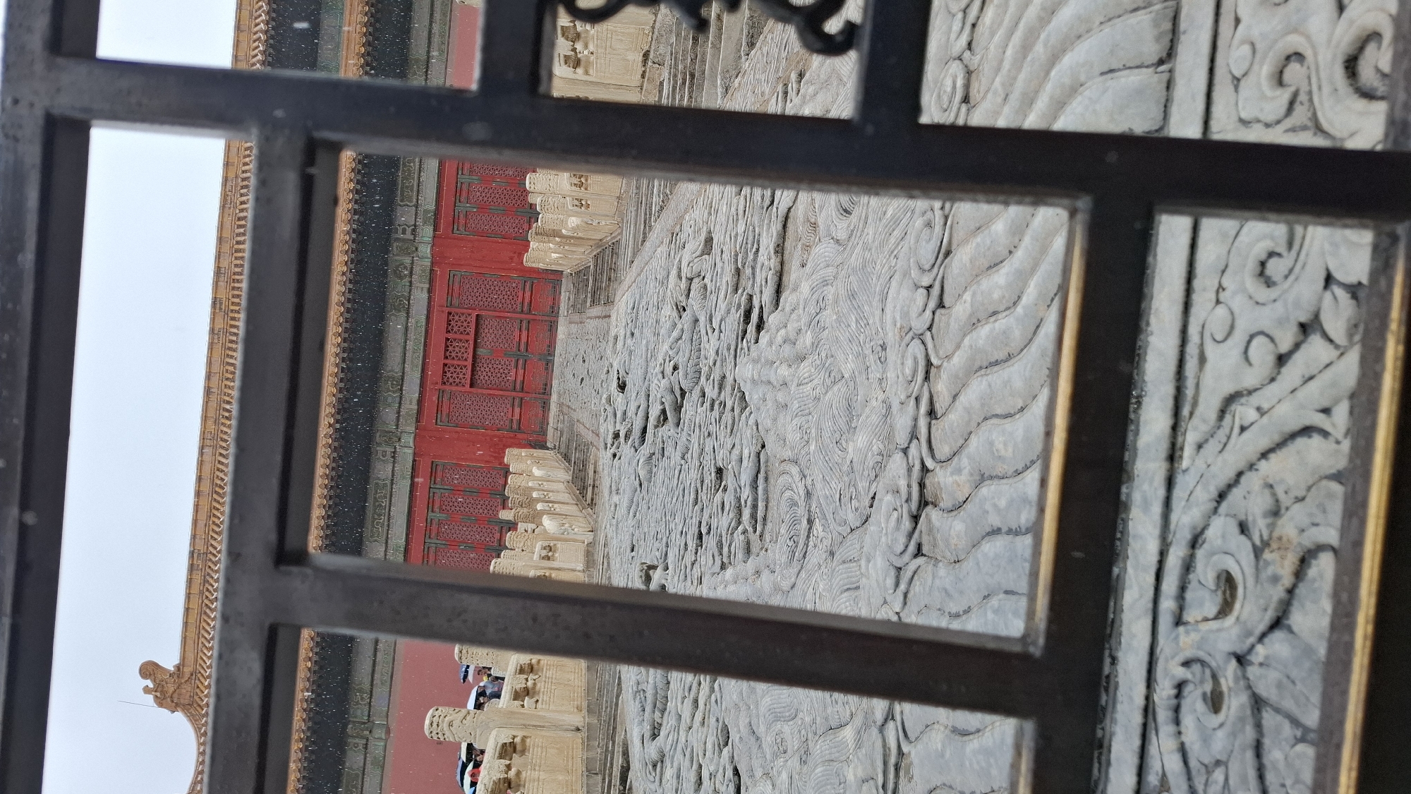 The image shows a view through a metal grate of architectural elements, likely in the city of Dongcheng District, China. Visible are a red building facade with patterned windows, a stone pathway carved with intricate designs, and sections of a roof with an ornate edge. There are also blurry images of people in the distance, standing at the base of the building.