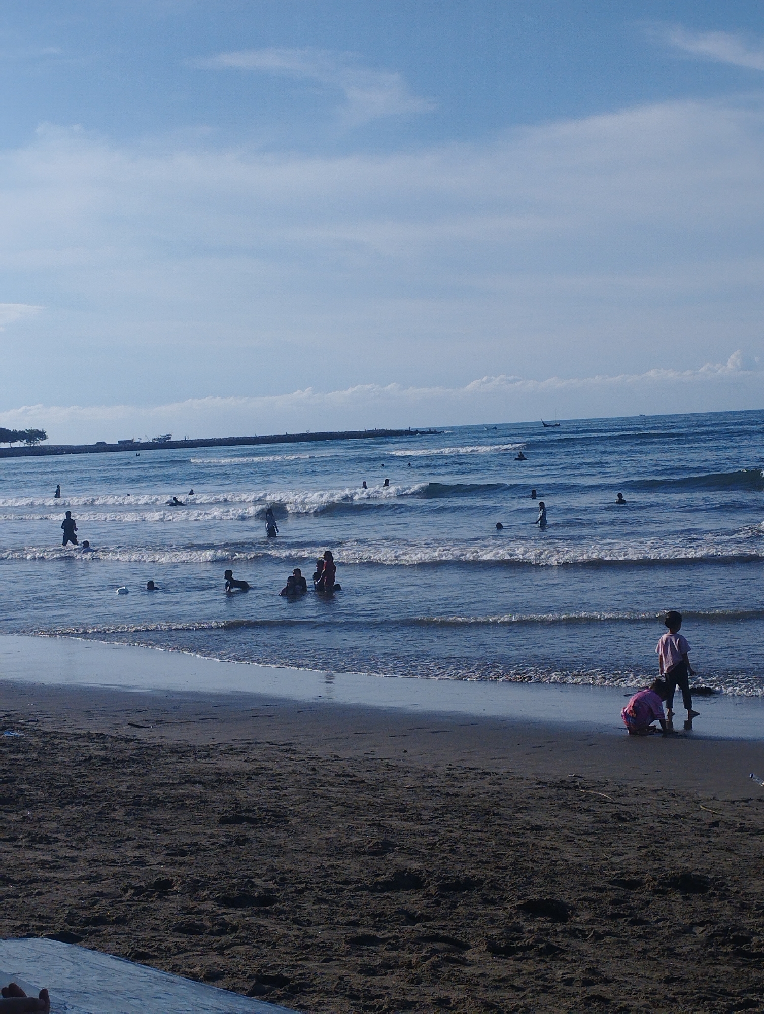 The image depicts a beach scene with multiple individuals present, possibly in Lhokseumawe, Indonesia. In the foreground, there is a dark sandy beach with two children. One child is crouched down, interacting with the sand, while the other is standing nearby, watching. Further into the scene, the ocean has small waves and a number of people are wading or swimming. Several groups of people are visible in the water, some standing and some submerged. There are also a couple of ducks in the water. The background features a breakwater or jetty extending out into the ocean. The sky is blue with scattered clouds. There is a visible vessel at a distance, with trees and low buildings on land to the left side of the scene.