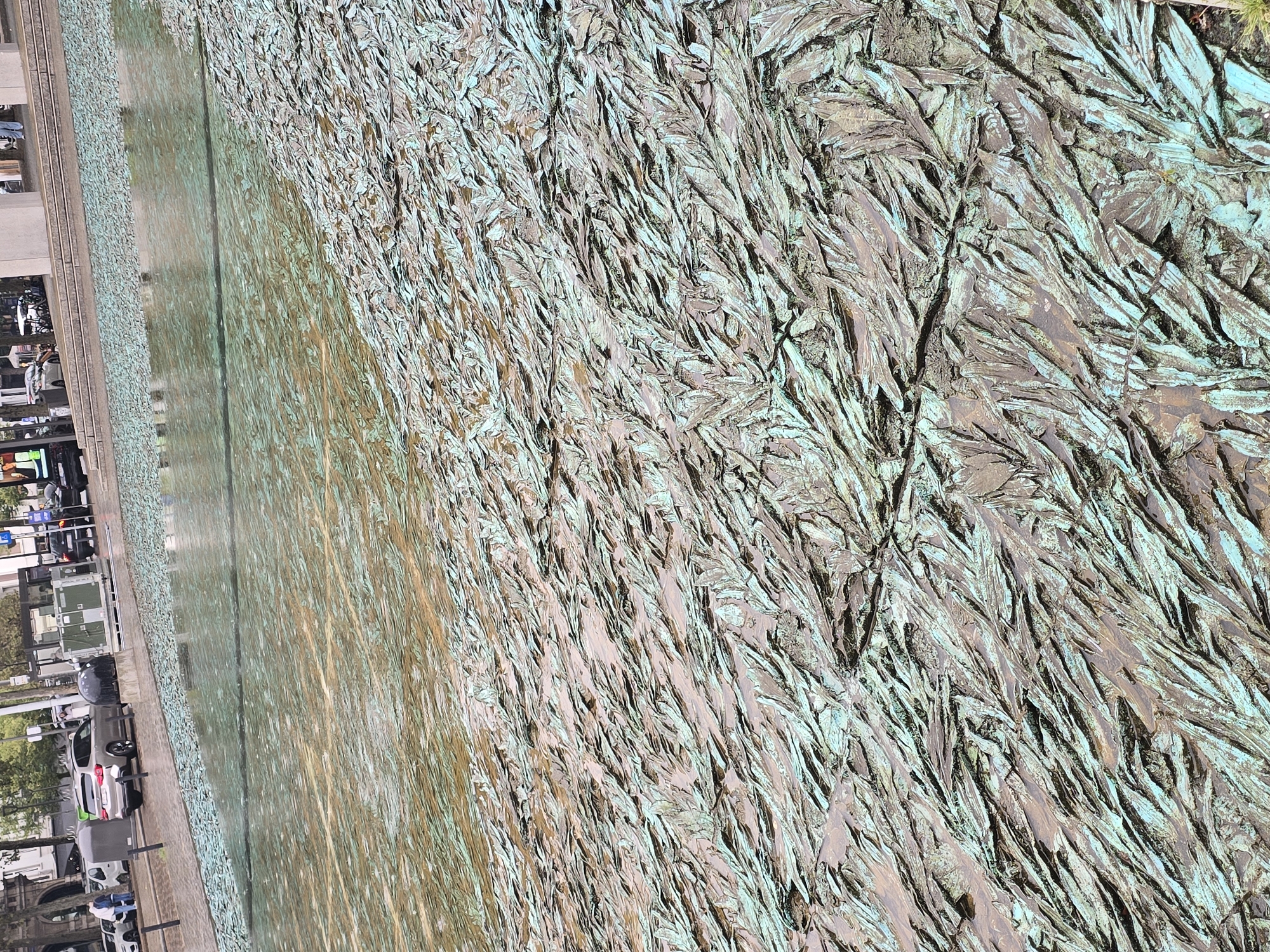 The image captures a section of the "Hoge Beeldekens," a public art installation in Antwerp, Belgium, designed by Luc Deleu. The artwork consists of a large, shallow water basin with a textured surface reminiscent of leaves or scales. The dominant colors are the muted green patina of the metal and the reflections of the sky and surrounding environment in the water.
In the background, the urban setting of Antwerp is visible. To the left, a silver SUV is parked near a building with large windows and an awning. Pedestrians are also present, including two individuals walking on a sidewalk. Across the street, there are more buildings, some with visible signage and architectural details, and bicycles are parked near a building. A portion of a black car is visible in the far background, and what appears to be a streetlamp or utility pole stands to the right of the water basin.
The foreground is dominated by the art installation itself. The textured surface, covered in shallow water, creates a dynamic visual effect. The water level is low, allowing the detailed texture of the metal to be clearly seen. The metallic surface exhibits a patina, with areas of greenish-blue oxidation and darker, tarnished metal. The arrangement of the textured elements creates a sense of depth and movement.