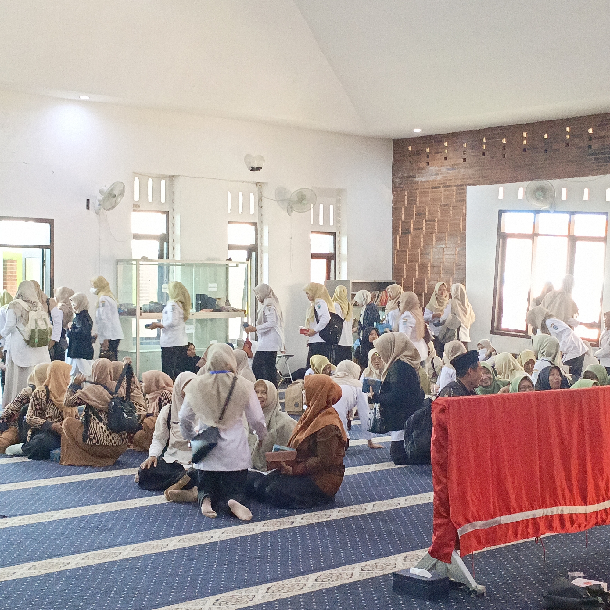 The image depicts an indoor gathering, likely at a mosque or religious educational facility in Salatiga, Indonesia. A large group of individuals, predominantly women wearing hijabs in various colors and patterns, are seated on a blue carpet with white and beige stripes. The room has high ceilings with white walls and exposed brickwork on one side, featuring small square openings near the ceiling for ventilation or light. Natural light streams in from windows on the right side of the image.
Several people are standing and interacting, some holding books or bags. One woman in a white hijab and black abaya is facing a glass display cabinet filled with items. Another woman in a brown hijab is holding a book. A man in a black cap and traditional attire is speaking to someone. Fans are mounted on the walls. A large red cloth with silver trim is prominently displayed in the foreground, supported by a stand. The overall atmosphere suggests a community event or educational session.