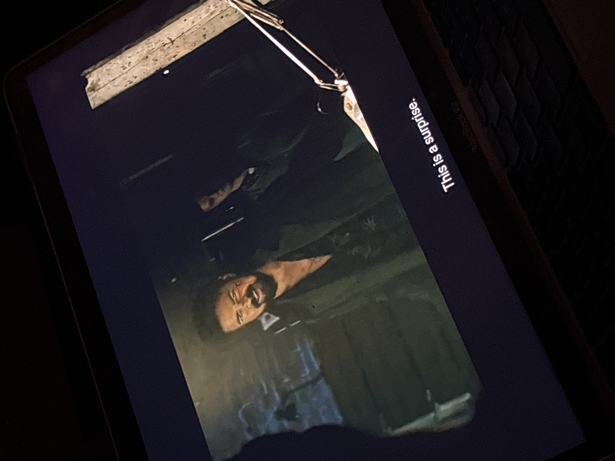 A slightly angled shot shows a tablet screen displaying a scene from a film or show. A man with a beard and a pained expression is visible. Text on the right reads "This is a surprise." Parts of a keyboard and some other ambient light are visible, indicating the shot was likely taken in a dimly lit indoor setting. The image does not contain any information to deduce the location.