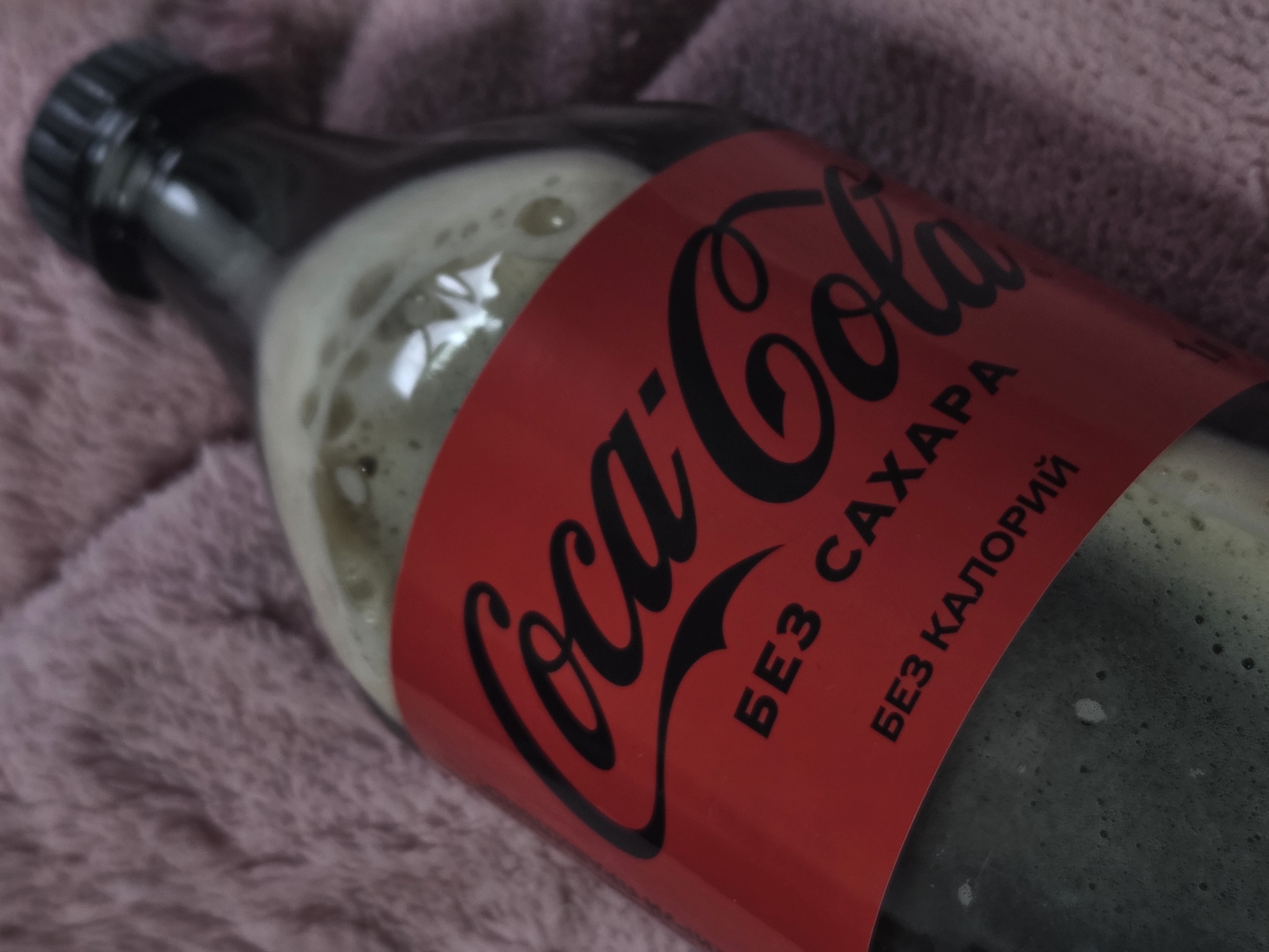 A close-up shot shows a bottle of Coca-Cola lying horizontally on a soft, textured, light purple or pink blanket. The bottle, dark in color, has a black screw-on cap visible on its left side. Inside, a dark, carbonated liquid is visible, topped with a significant layer of light-colored foam, suggesting the drink might have been recently opened or shaken.
The prominent red label on the bottle features the iconic black "Coca-Cola" script logo. Below the main logo, two lines of text are printed in black Cyrillic characters: "БЕЗ САХАРА" (SUGAR FREE) and "БЕЗ КАЛОРИЙ" (CALORIE FREE), indicating that it is a sugar-free variant of the beverage. The soft, fuzzy texture of the blanket provides a contrasting background to the smooth surface of the bottle and its label. The lighting is diffuse, highlighting the details of the foam, the bottle's reflections, and the fabric's texture.