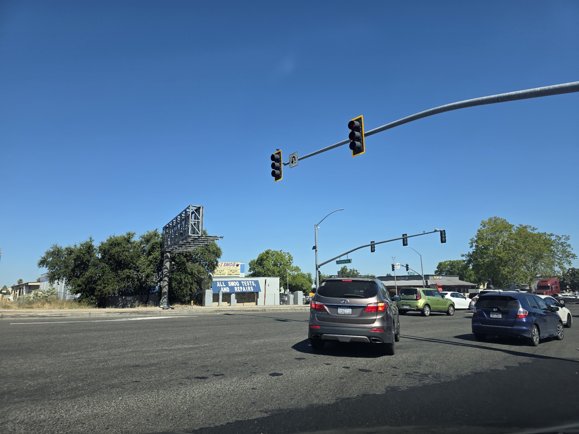 The image depicts a sunny daytime scene at a busy intersection in West Sacramento, California. The sky is a clear, bright blue, suggesting fair weather. The foreground is dominated by an asphalt road, marked with white lines, indicating multiple lanes of traffic.
Several vehicles are present, including a gray SUV in the right lane, a blue hatchback behind it, and a lime green car in the left lane, all seemingly stopped at a red light. Further down the road, other cars and a red semi-truck are visible, suggesting ongoing traffic flow.
Above the intersection, traffic lights hang from a large metal pole that spans across the road. One set of traffic lights shows red for all bulbs, while another set, further to the left, also shows red. A street sign indicates "Carmichael Ave."
To the left of the intersection, a large industrial-style billboard structure stands tall. Adjacent to it is a building with a blue banner that reads: "ALL SMOG TESTS, AND REPAIRS." The building appears to be a business, possibly a smog testing and repair shop. The overall scene conveys the everyday hustle and bustle of a suburban or commercial area. There are no people visible in the image.