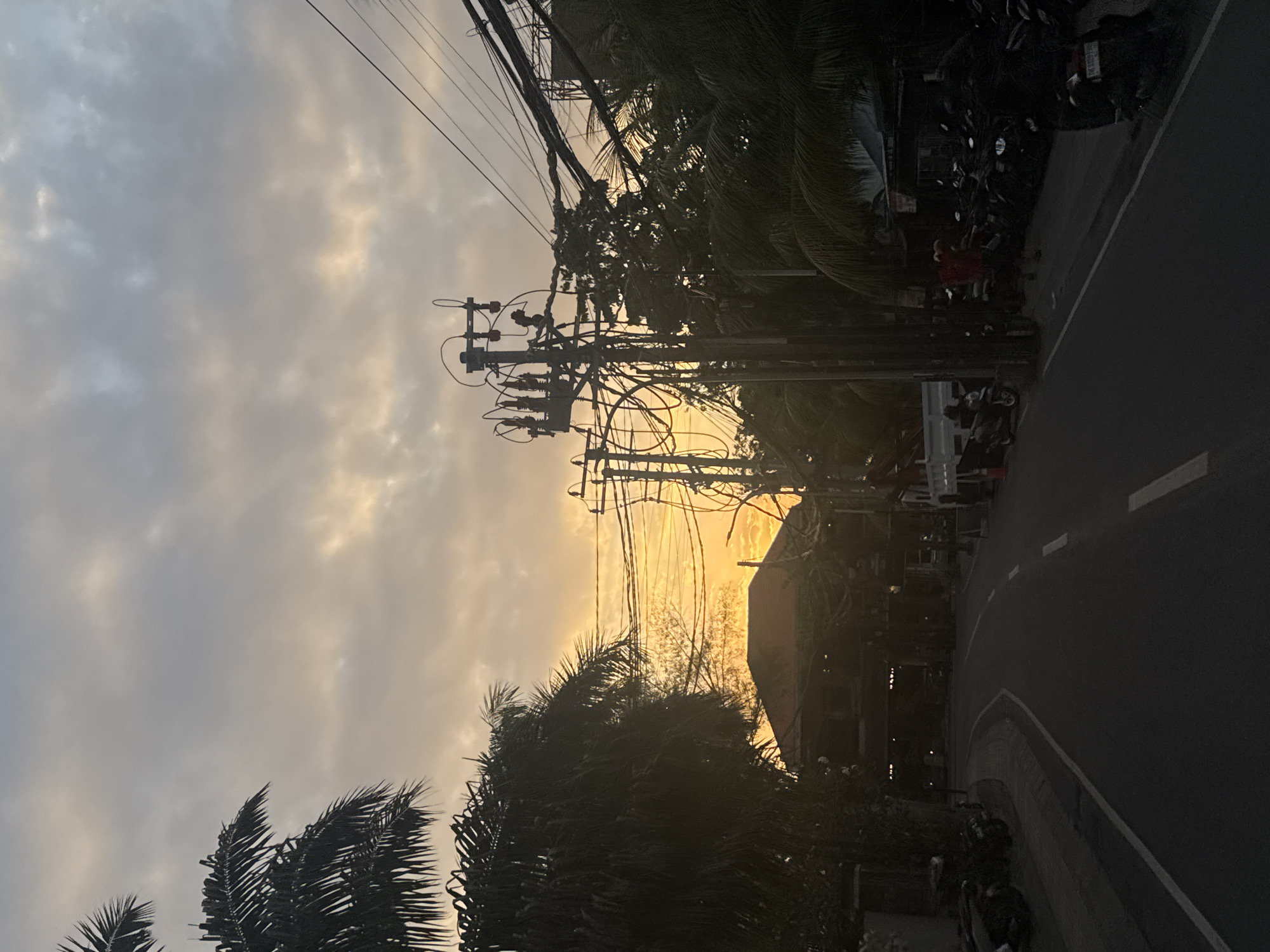 This image captures a street scene in North Kuta, Indonesia, during either sunrise or sunset. A two-lane road curves towards the right, bordered by palm trees and various structures. A dense network of utility poles and electrical wires dominates the mid-ground, crisscrossing the frame. On the left side of the road, parked motorbikes are visible. Further down the road, on the right, another group of parked motorbikes lines the sidewalk, with a few individuals standing nearby. A white truck is parked on the left side of the road. The sky is overcast with bands of orange and yellow light breaking through the clouds, suggesting the presence of the sun below the horizon. The overall atmosphere is subdued, with the lighting casting long shadows and obscuring some details.