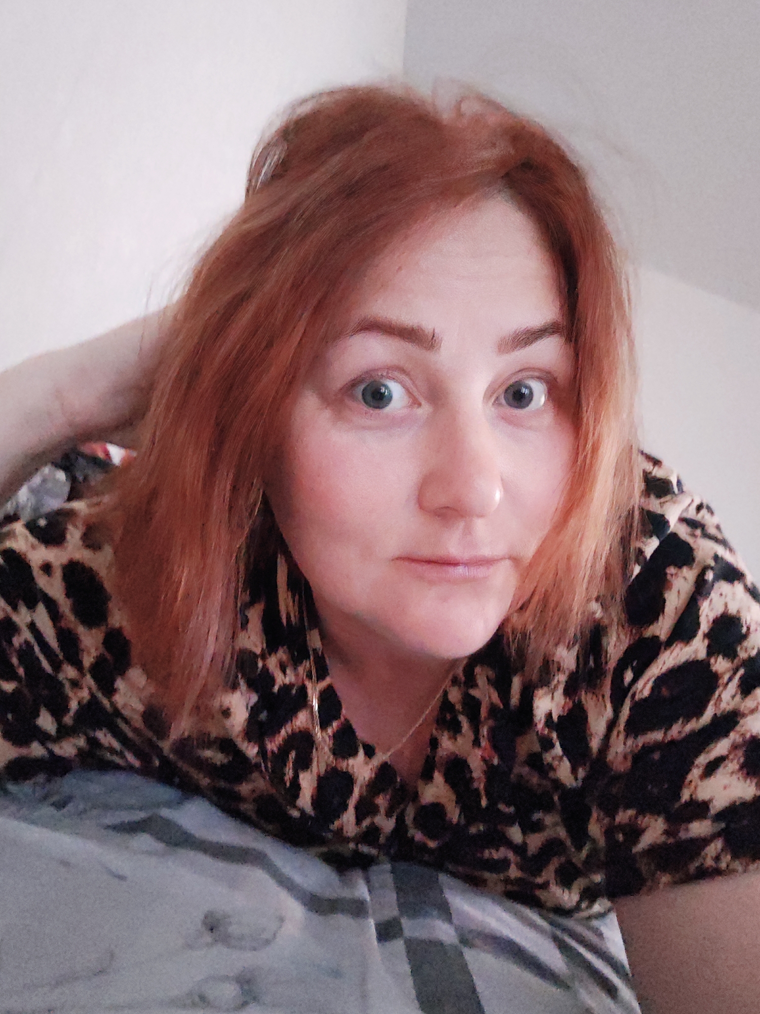 A woman with red hair, wearing a leopard print top, is looking at the camera. She is indoors, lying down on a grey and white patterned surface, possibly a bed. One hand is raised near her head. She appears to have a nose ring and gold earrings. The background is a plain white wall. The location in Markovskii, Russia is not evident from the image.