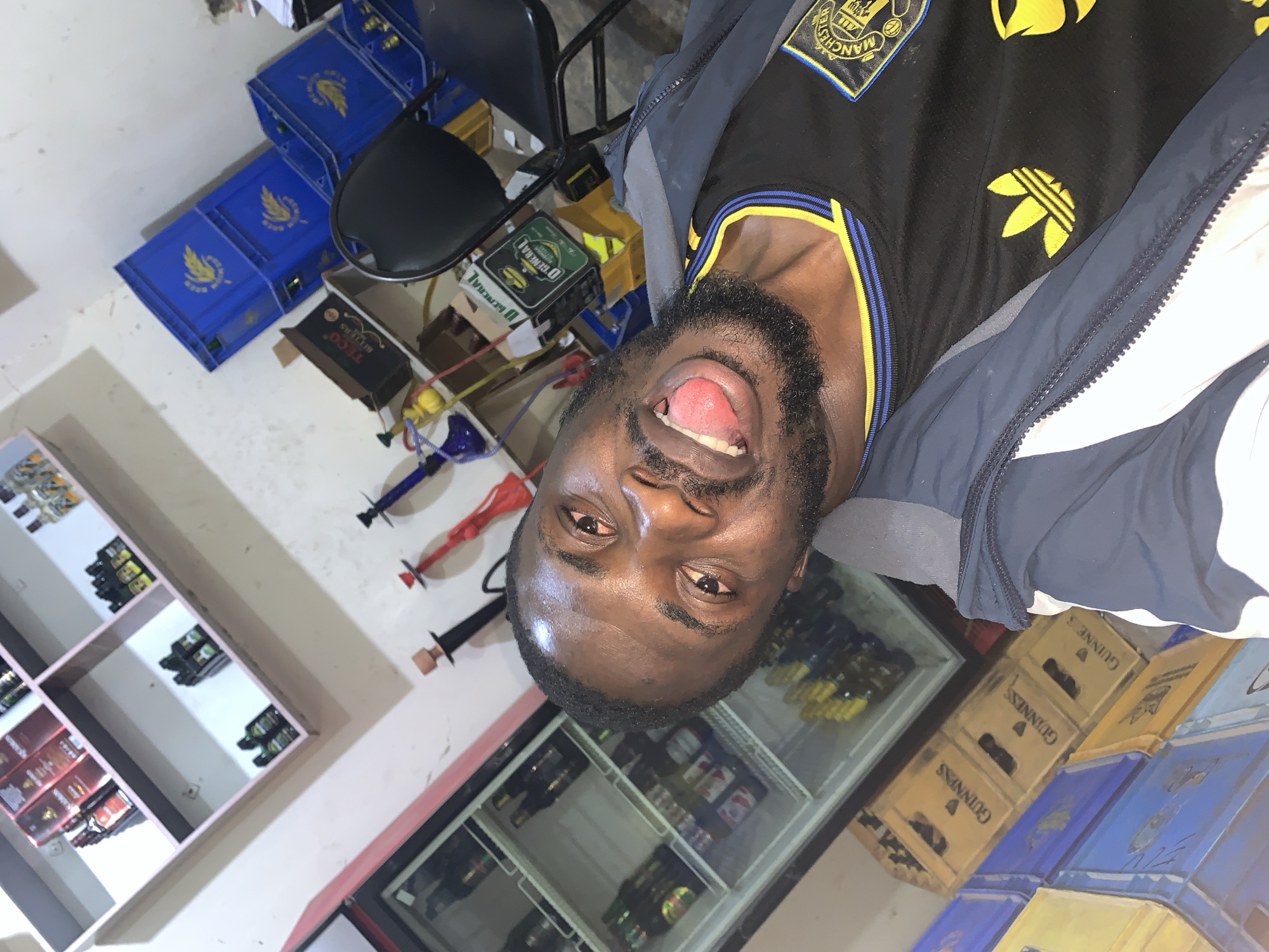 A man is pictured in a room that appears to be a shop or bar, possibly located in New Stadium, Nigeria. The man is in the foreground, facing the camera, with his mouth open and tongue sticking out. He is wearing a black and yellow Adidas shirt with a Manchester United crest. Behind him, shelves are stocked with various bottles, and to his right, blue crates are stacked. A refrigerator is visible to his left, filled with bottles. The environment suggests a commercial setting, likely selling beverages.