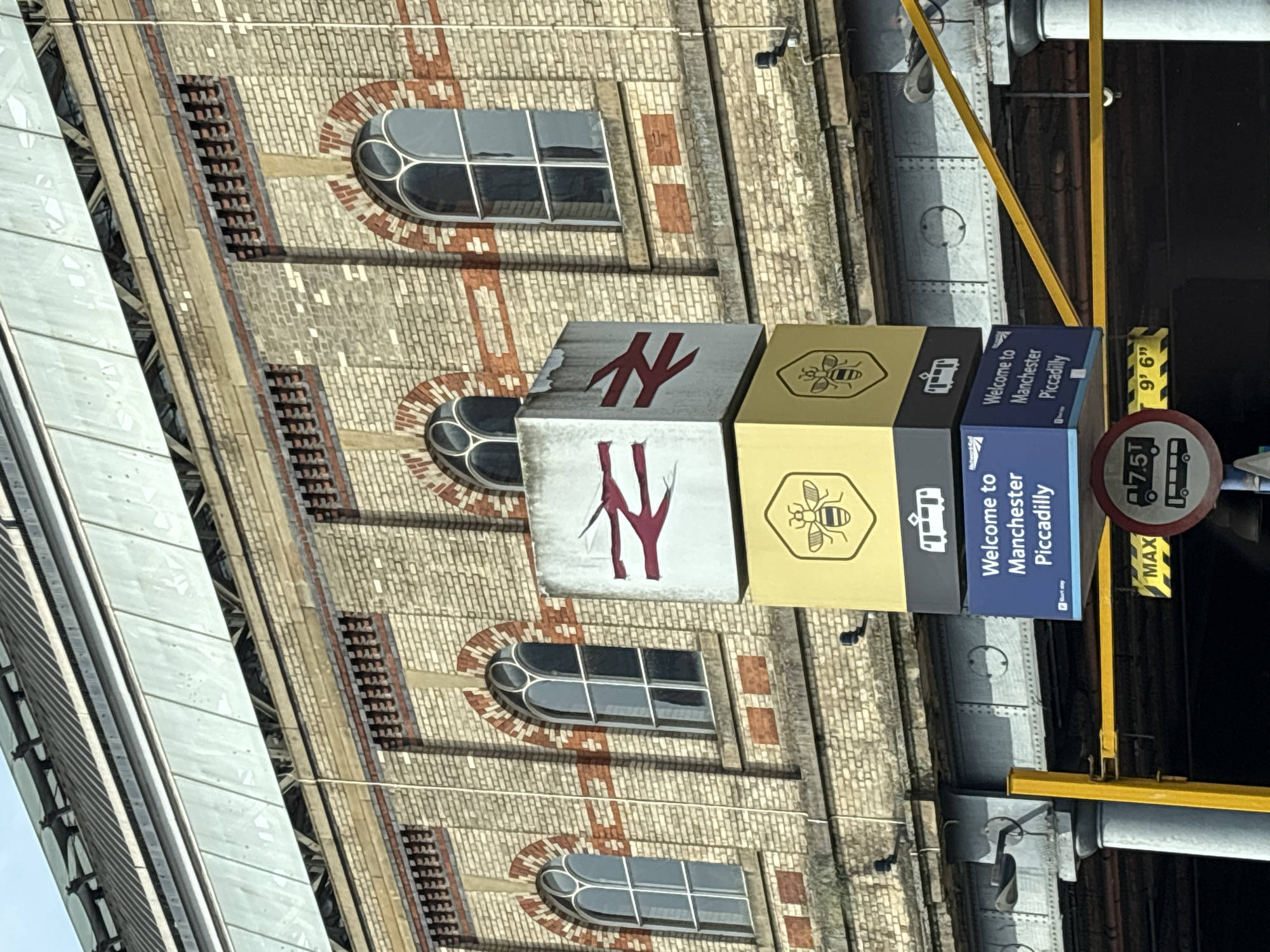 The image shows a vertically oriented view of a signpost at Manchester Piccadilly train station in Manchester, United Kingdom. The signpost is comprised of stacked cubes with the National Rail logo on the top cubes, a bee symbol (a symbol of Manchester) on the next cube down, and "Welcome to Manchester Piccadilly" on the bottom cube. Behind the sign is the station's brick exterior with arched windows. A weight limit sign is also visible next to the signpost. The image is rotated.