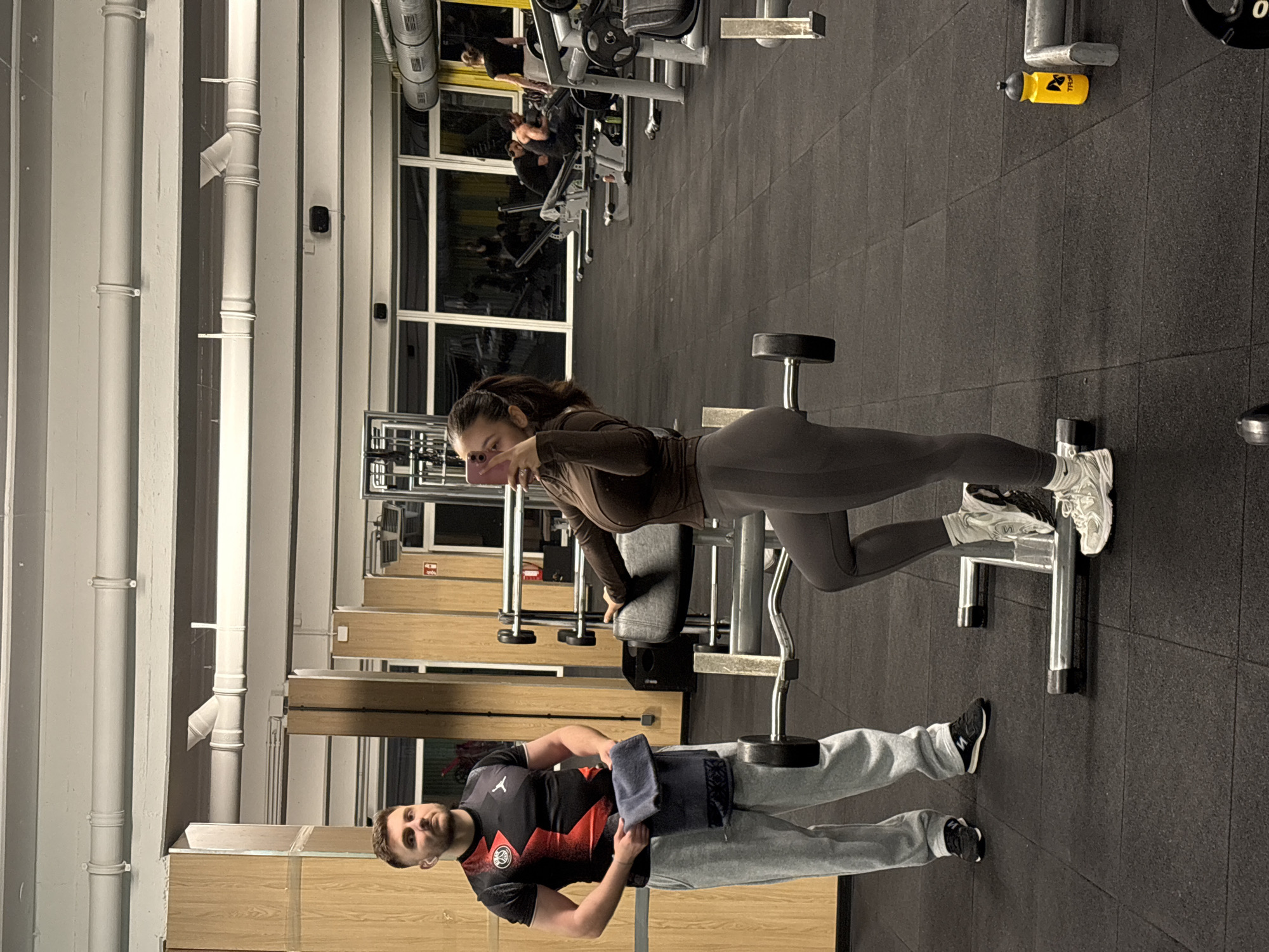 The image captures a gym scene in Mönchengladbach, Germany. A woman in a brown long-sleeved top and grey leggings is using a weight machine. She's holding a phone and making a peace sign. A man in a black and red Paris Saint-Germain jersey and grey sweatpants stands near her with a grey towel. Gym equipment is visible in the background, including treadmills and other weight machines. The floor is black rubber. Pipes run along the ceiling, and windows provide natural light.
