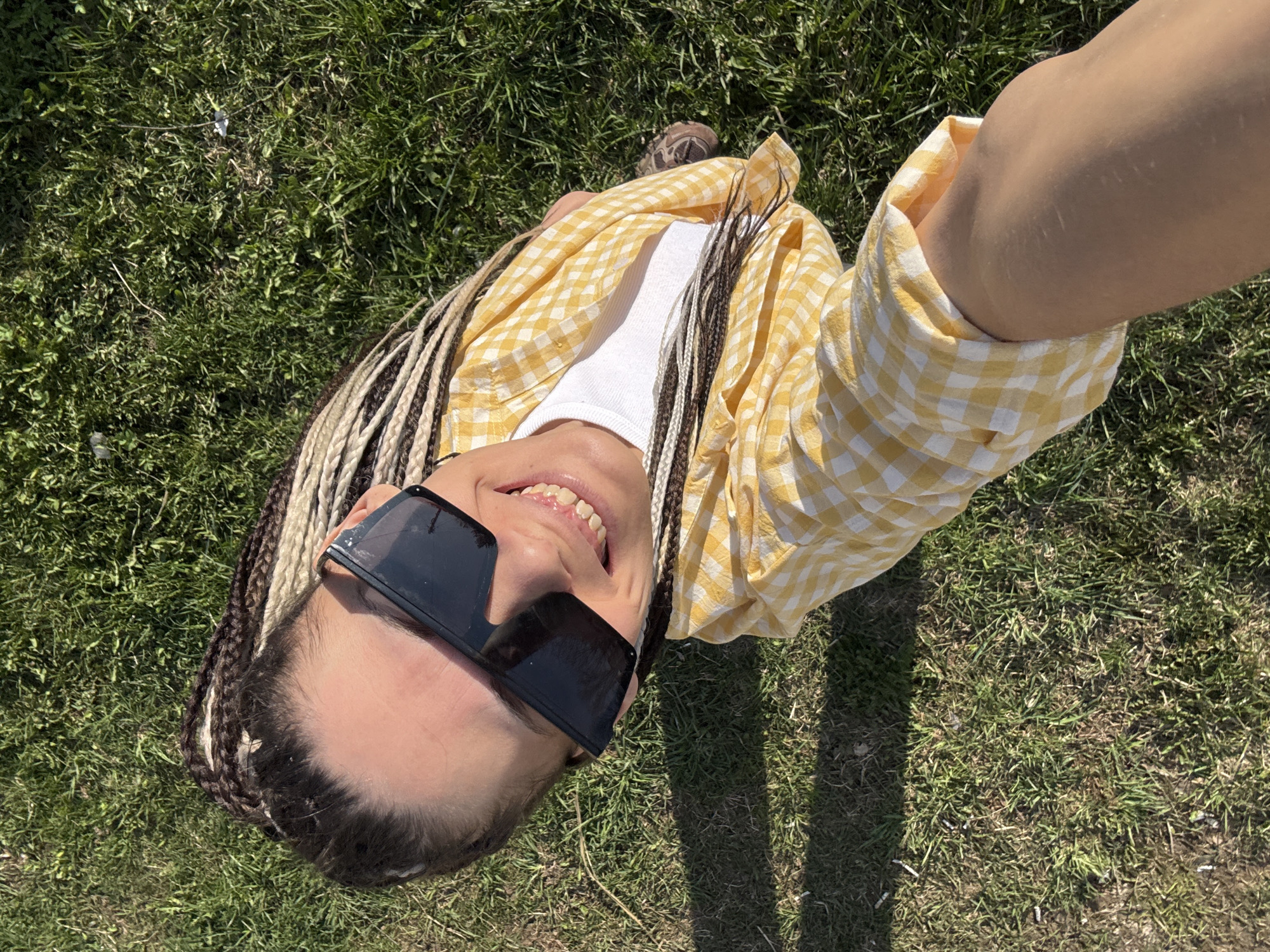 The image shows a person lying on grass, possibly in Fedorino, Russia, although this cannot be confirmed from the visual data alone. The person is wearing sunglasses and a yellow and white checkered shirt over a white undershirt. They have braided hair with a mix of brown and blonde strands. The camera angle is from above, possibly a selfie taken with an extended arm. The background is primarily green grass. There is a shoe visible in the top-left corner. The grass has some light patches. The person is smiling.