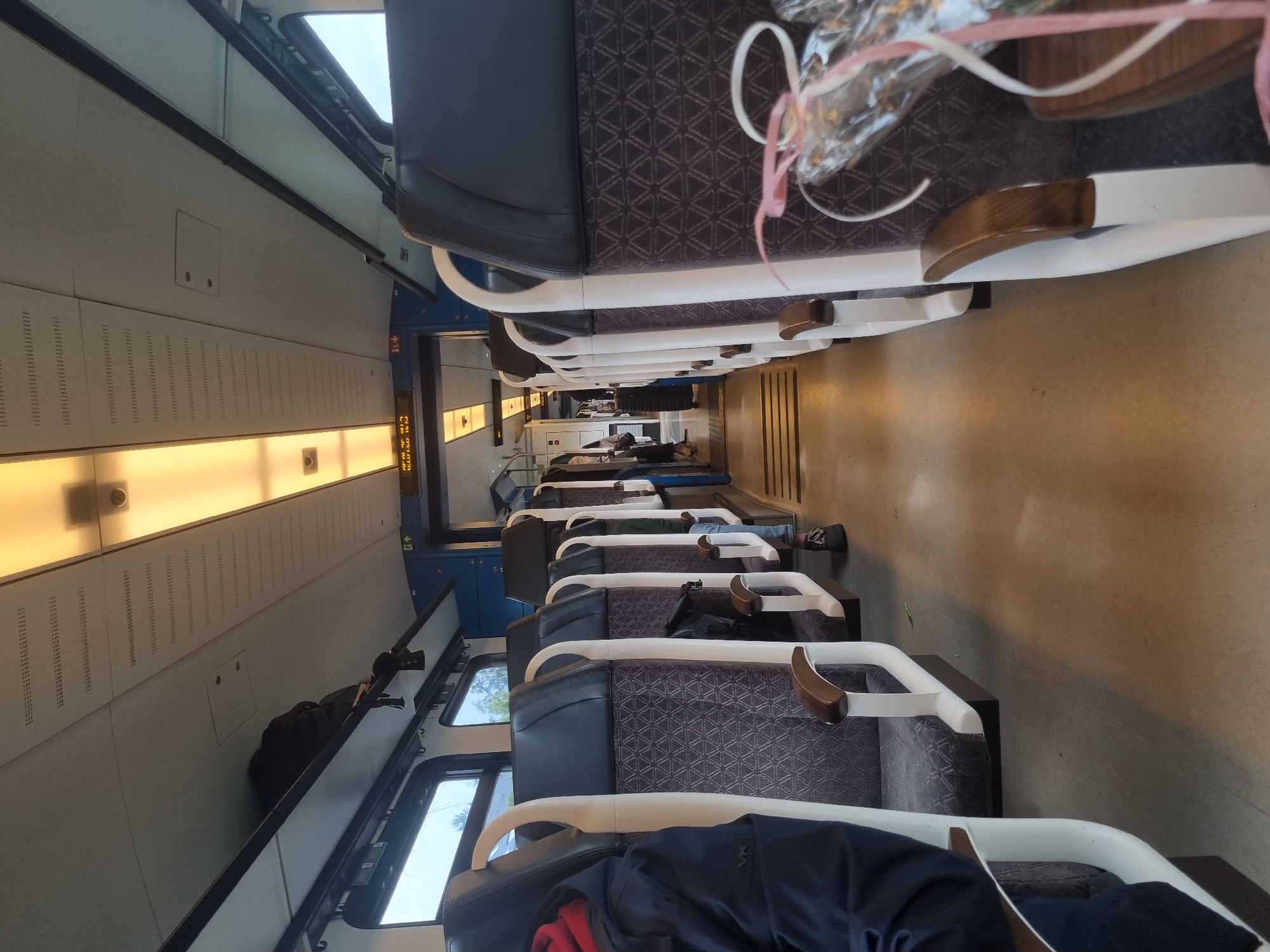 The image depicts the interior of a train car, likely in Cochem, Germany. Rows of seats with a dark blue and gray geometric pattern extend into the distance. Some seats are occupied by passengers, with a couple visible. Bags and clothing are draped over some of the seats. Windows are visible on the left side of the frame. An illuminated display panel is on the wall. The overall scene suggests a journey in progress.