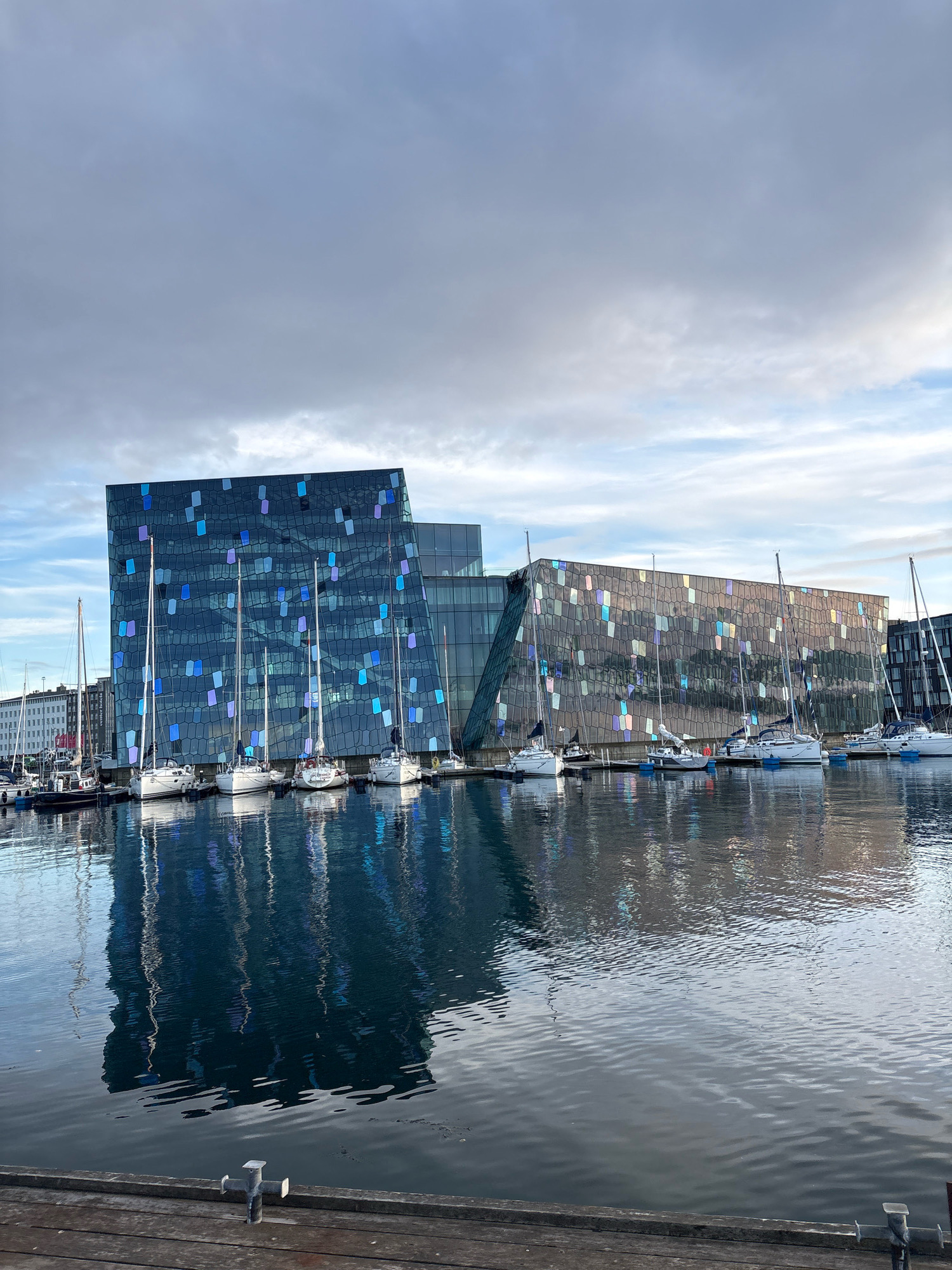 The image shows the Harpa Concert Hall and Conference Centre in Reykjavik, Iceland. The modern building features a distinctive glass facade with geometric patterns. In front of the building, several sailboats are docked in a harbor. The water reflects the building and the boats. The sky is cloudy, with hints of blue peeking through. In the foreground, a wooden dock with a metal cleat is visible. There are no people visible in the image.