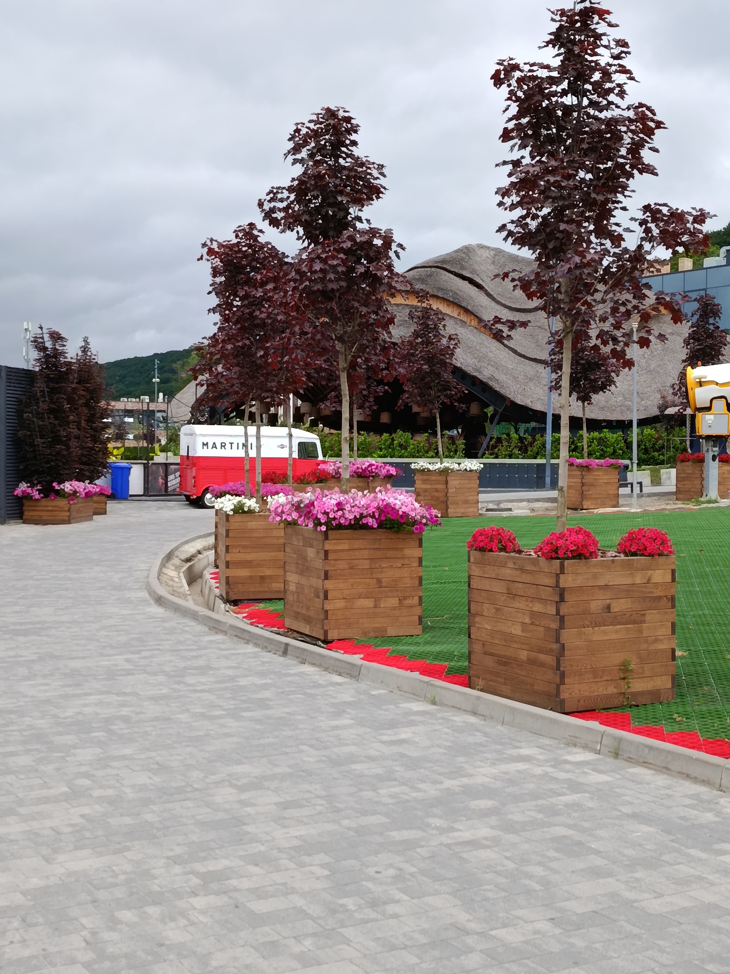The image depicts an outdoor landscaped area in Vynnyky, Ukraine, under an overcast sky. A light grey paver pathway, composed of rectangular blocks in a staggered pattern, occupies the left and bottom portions of the foreground, curving towards the right. This path is bordered by a grey concrete curb, adjacent to which a red, segmented ground-level strip lies on a green artificial turf or textured ground covering.
Numerous wooden rectangular planters, constructed from horizontally stacked planks, are arranged on this green surface. The planters in the immediate foreground contain clusters of vibrant red flowers. Further back, other planters display pink and white flowers. Several deciduous trees with dark reddish-purple leaves are strategically planted throughout the green area, some reaching considerable height.
In the mid-ground, a vintage-style mobile trailer, painted red with a white roof, is parked on a paved surface to the left of the pathway. The word "MARTINI" is displayed in black capital letters on its white side. A blue cylindrical waste bin is positioned nearby. Behind the trees, a distinctive building with a multi-sectioned, light brown thatched roof and visible wooden construction is a prominent feature. To its right, a section of a modern building with a dark glass facade is visible. On the far right, a yellow, tall, and somewhat rounded structure, possibly an observation device or a utility fixture, stands partially obscured by a tree.
The background reveals undulating green hills or mountains beneath a uniformly grey, cloudy sky. No individuals or animals are present in the depicted scene.