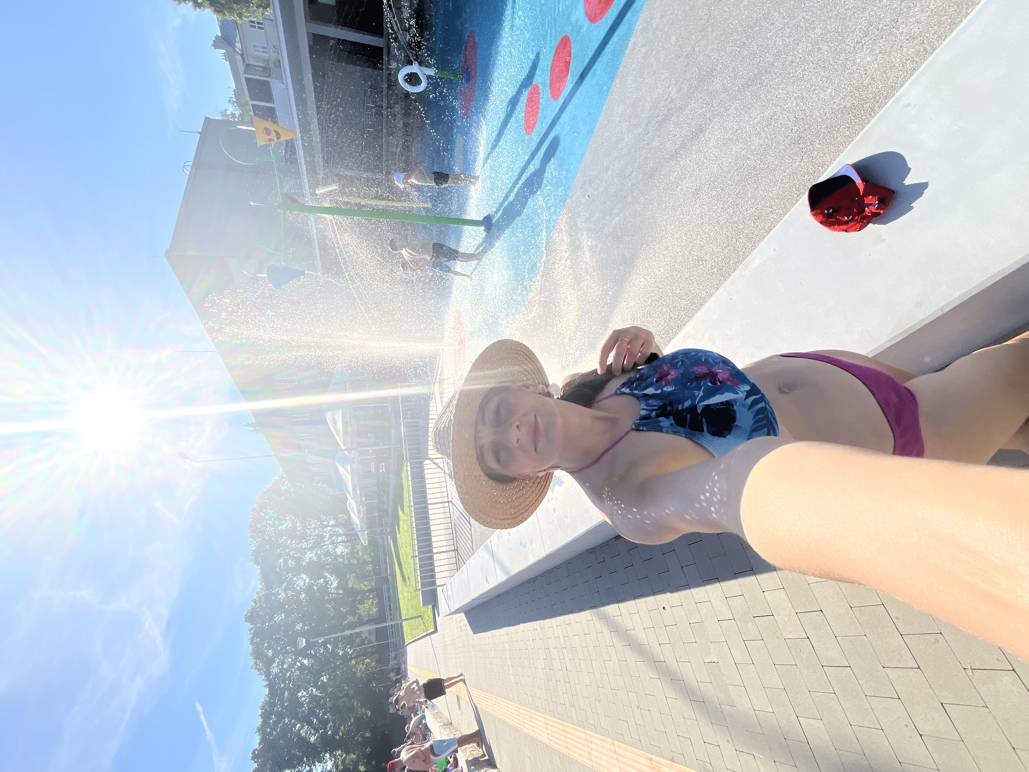 An outdoor, sun-drenched, wide-angle selfie taken at a public water play area. The foreground features a woman wearing a straw hat and a floral bikini, holding the camera with her right arm extended. She is looking towards the camera with a slight smile. To her right, on a white ledge, is a red and black baseball cap. The background depicts a blue water play area with a green fountain structure in the center, from which water sprays. Several children are visible playing in the water. The ground is paved with light-colored interlocking bricks, and there are trees and buildings in the distance. The sun is shining brightly, creating lens flare and casting shadows.