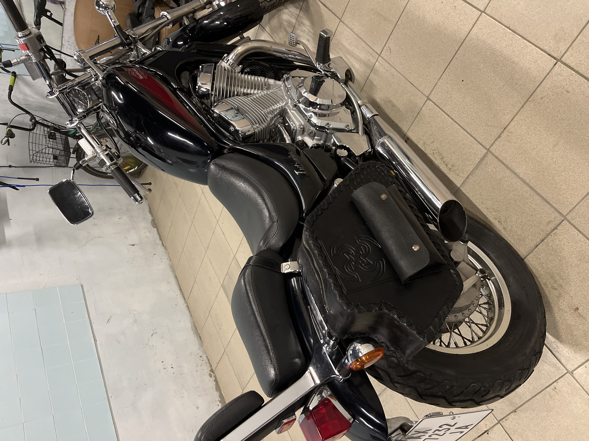 This is a medium-angle, eye-level shot of a black Suzuki Intruder motorcycle parked indoors, likely in a garage or storage unit. The motorcycle is oriented to the right, with its rear tire and saddlebag prominently featured in the foreground. The motorcycle's chrome engine is visible, as is its black leather saddlebag with embossed detailing. The background shows a tiled floor and a wall with a light blue tiled section and some white plaster. A bicycle with a basket is visible in the background, leaning against the wall. The license plate of the motorcycle, partially visible, reads "AX 7232 JA". The scene is static, with no people or active interactions. The lighting appears to be artificial, casting soft shadows. The specific location within the city of Myrhorod, Ukraine, cannot be determined from the visual information provided.